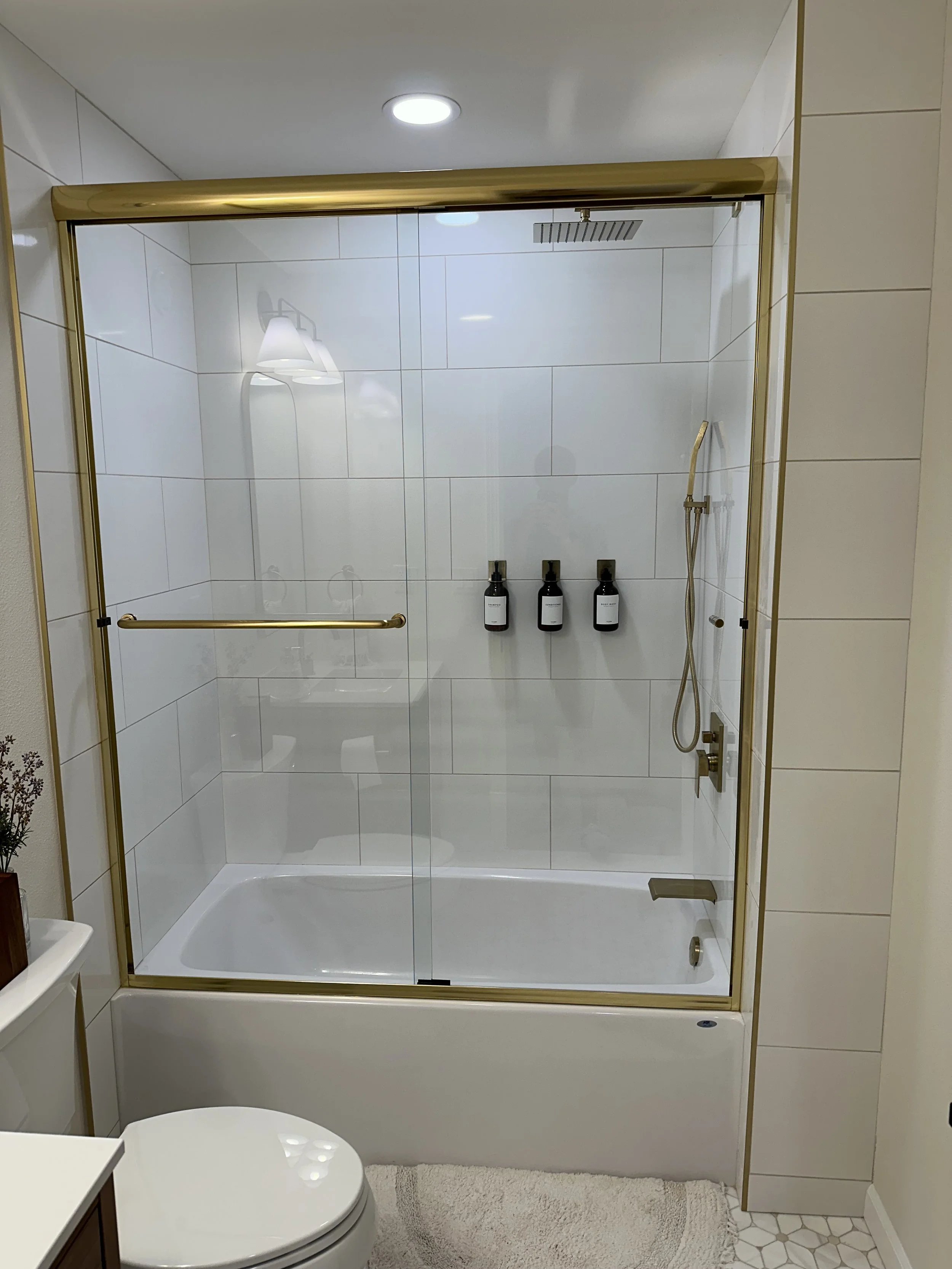 A modern glass shower enclosure with gold-colored trim and a sliding door, containing a rainfall showerhead, a handheld showerhead, and three black bottles of shampoo or body wash mounted on the wall, in a white tiled bathroom with a beige rug and a white toilet.