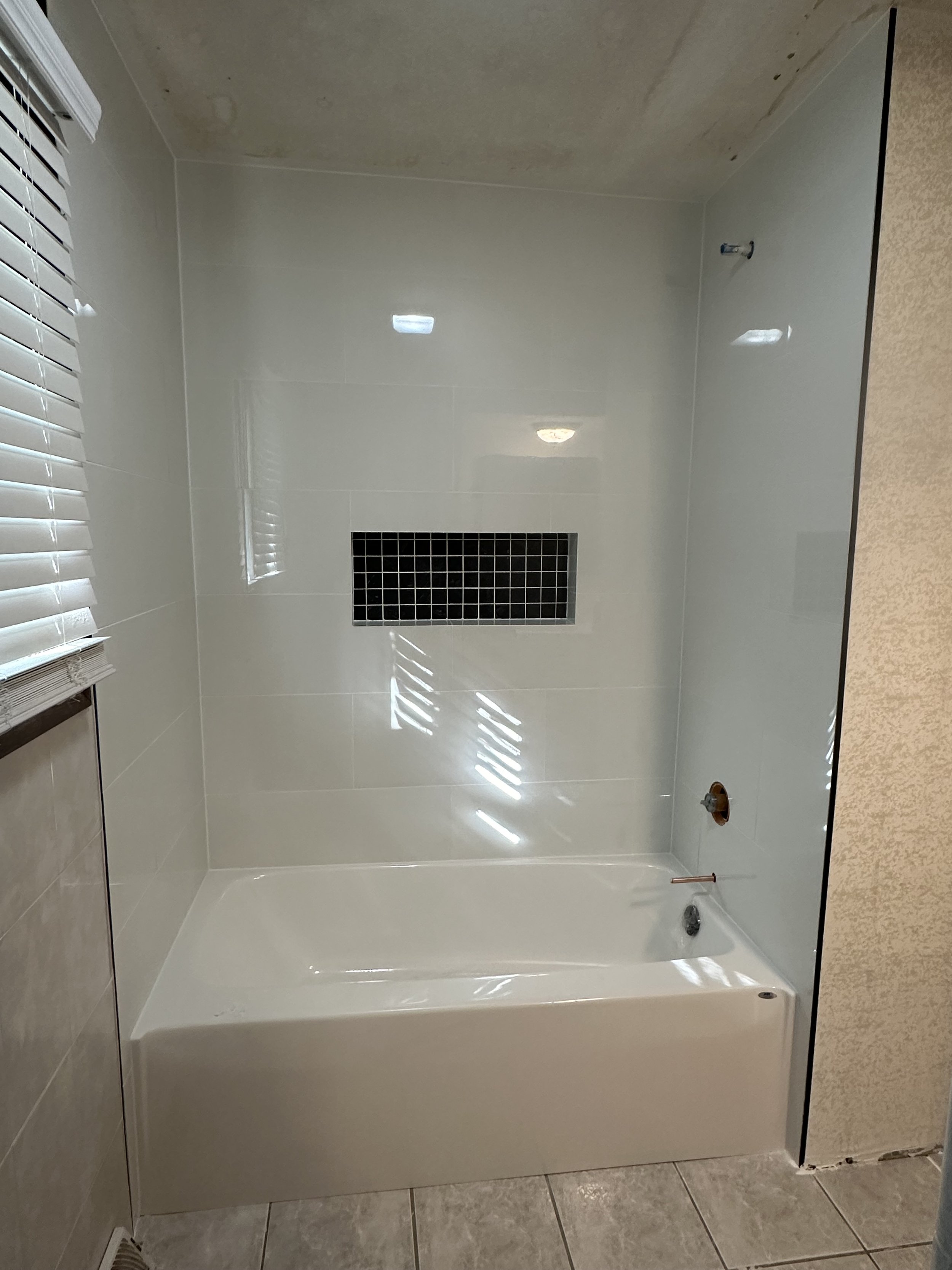 Empty white bathtub with a black drain cover, a copper pipe, and a faucet, in a bathroom with beige tiled floor and a white tiled wall. There is a window with white blinds and a plumbing vent.