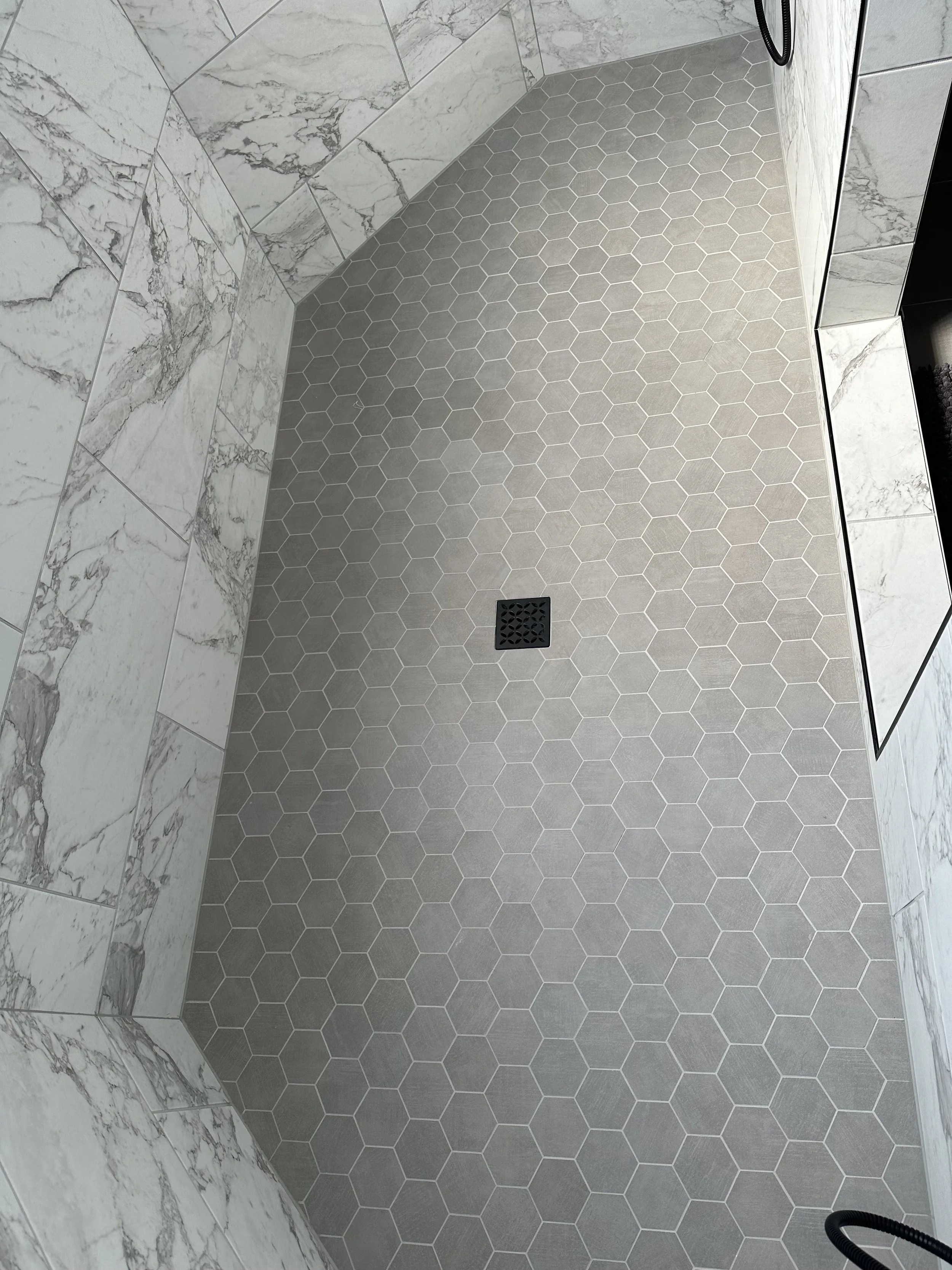 A bathroom shower with white marble wall tiles and gray hexagon floor tiles, including a drain in the center and a black hose on the wall.