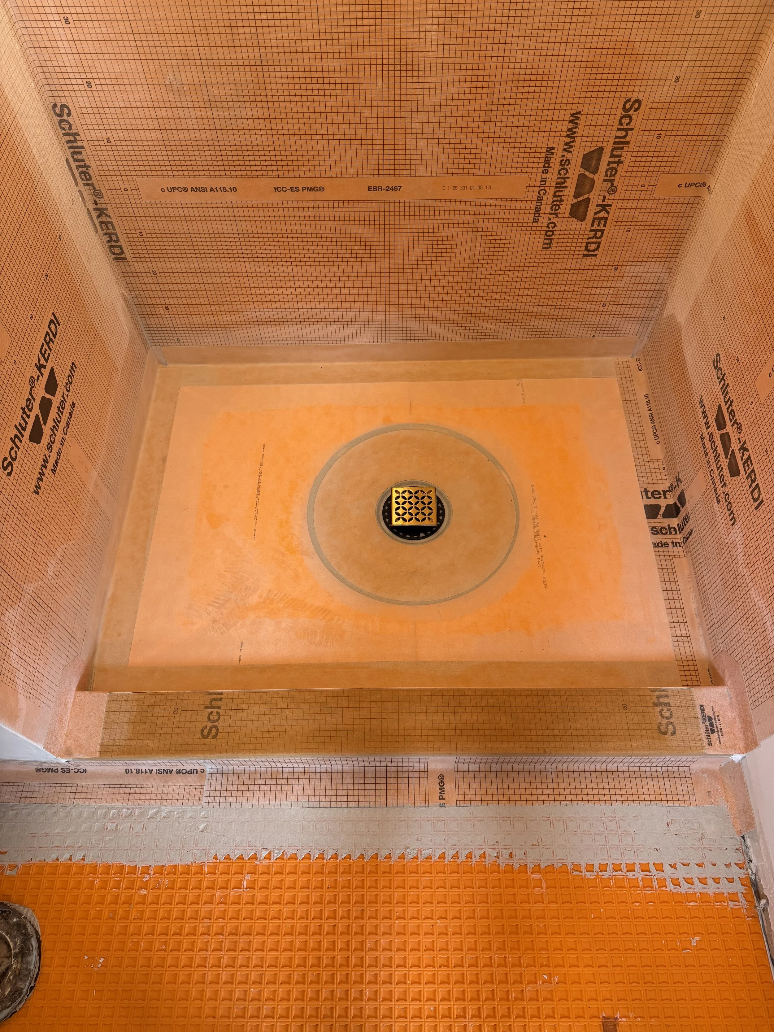 The photo shows a corner of a bathroom under construction with orange waterproofing membranes on the walls and floor, along with a drain on the floor.