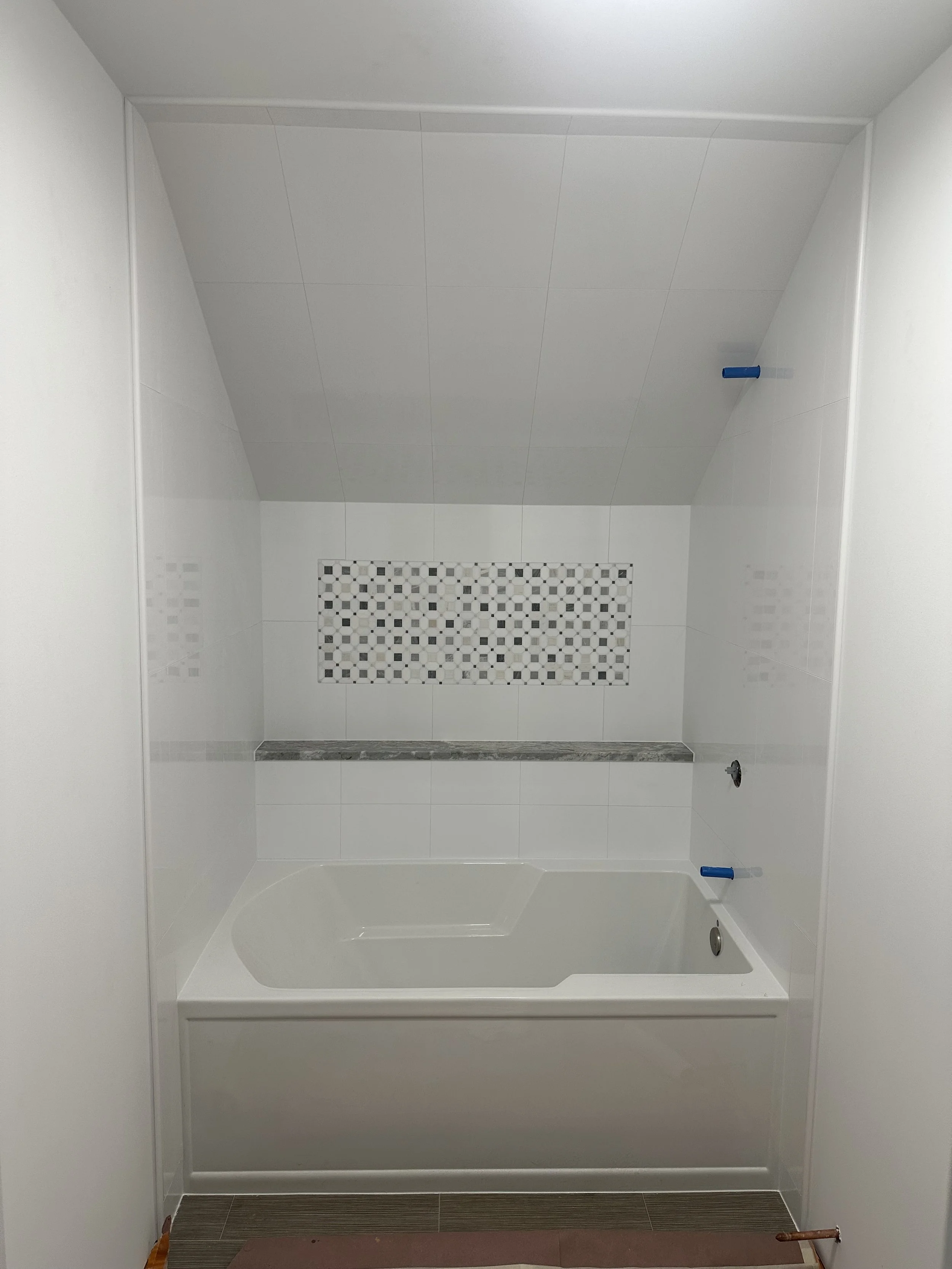 An unfinished shower stall with white walls and a tiled wall niche, bathroom fixtures not installed yet, with blue plumbing caps visible.