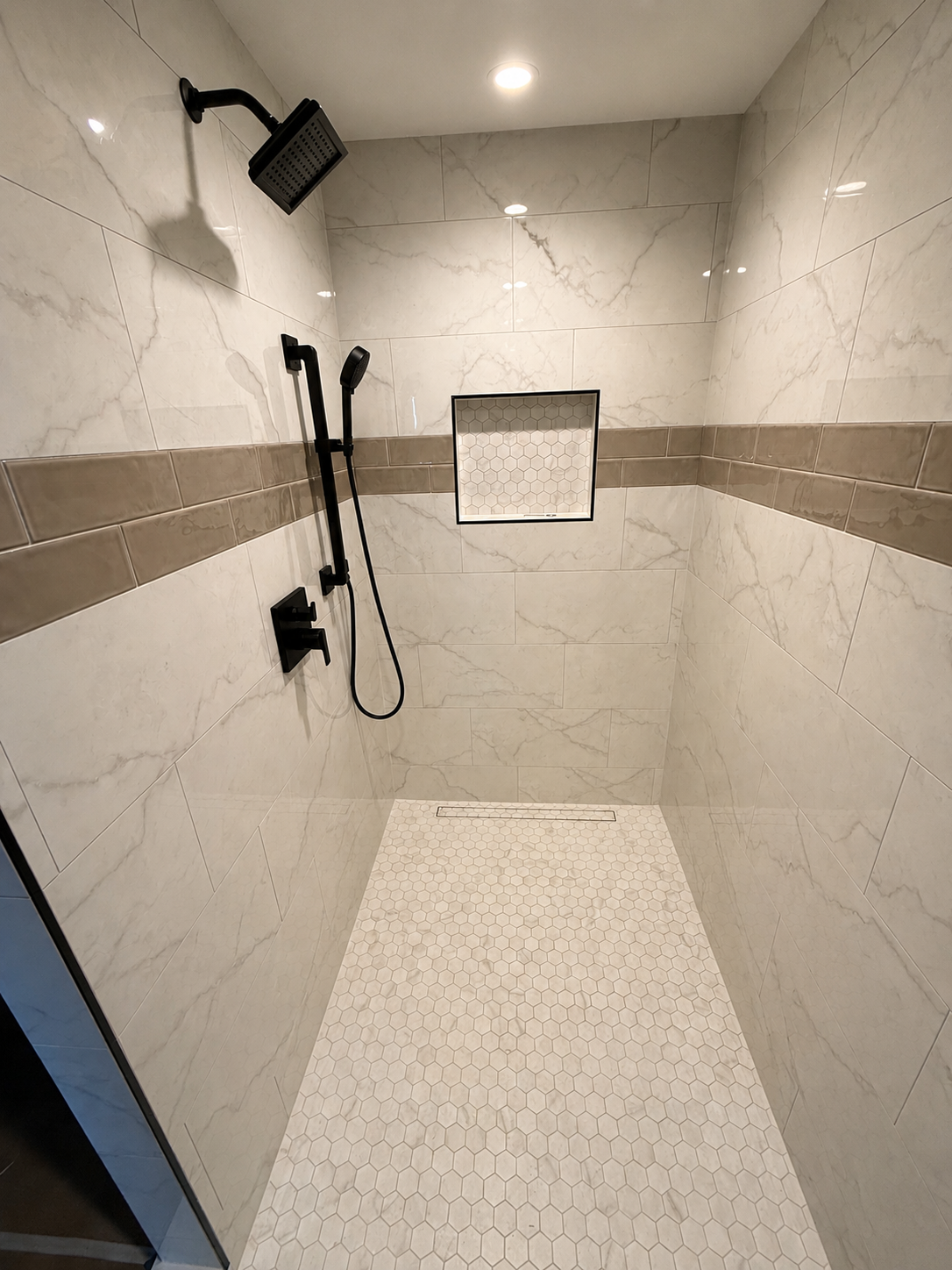 Empty walk-in shower with beige and white marble tiles, black showerhead and controls, small square mosaic tile inset, and a linear drain.