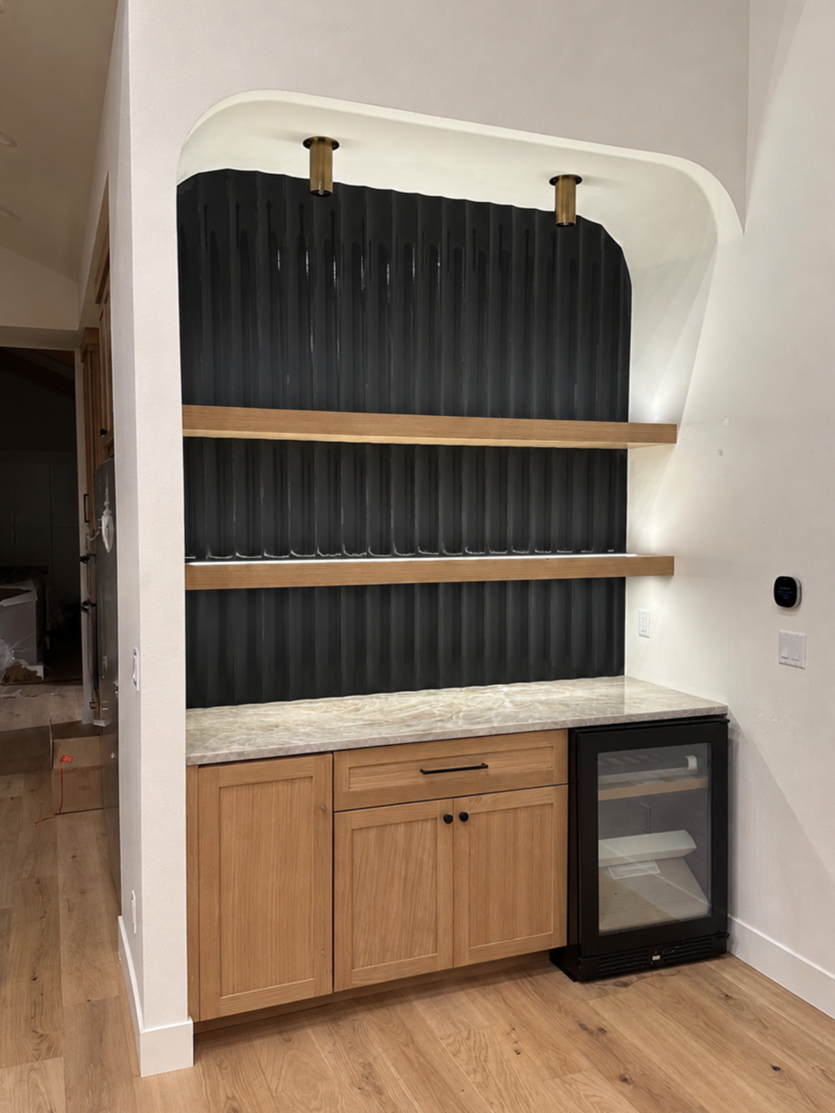 Kitchen bar area with wooden cabinets, marble countertop, black corrugated metal wall, two wooden shelves, a small wine cooler, and modern wall lighting.