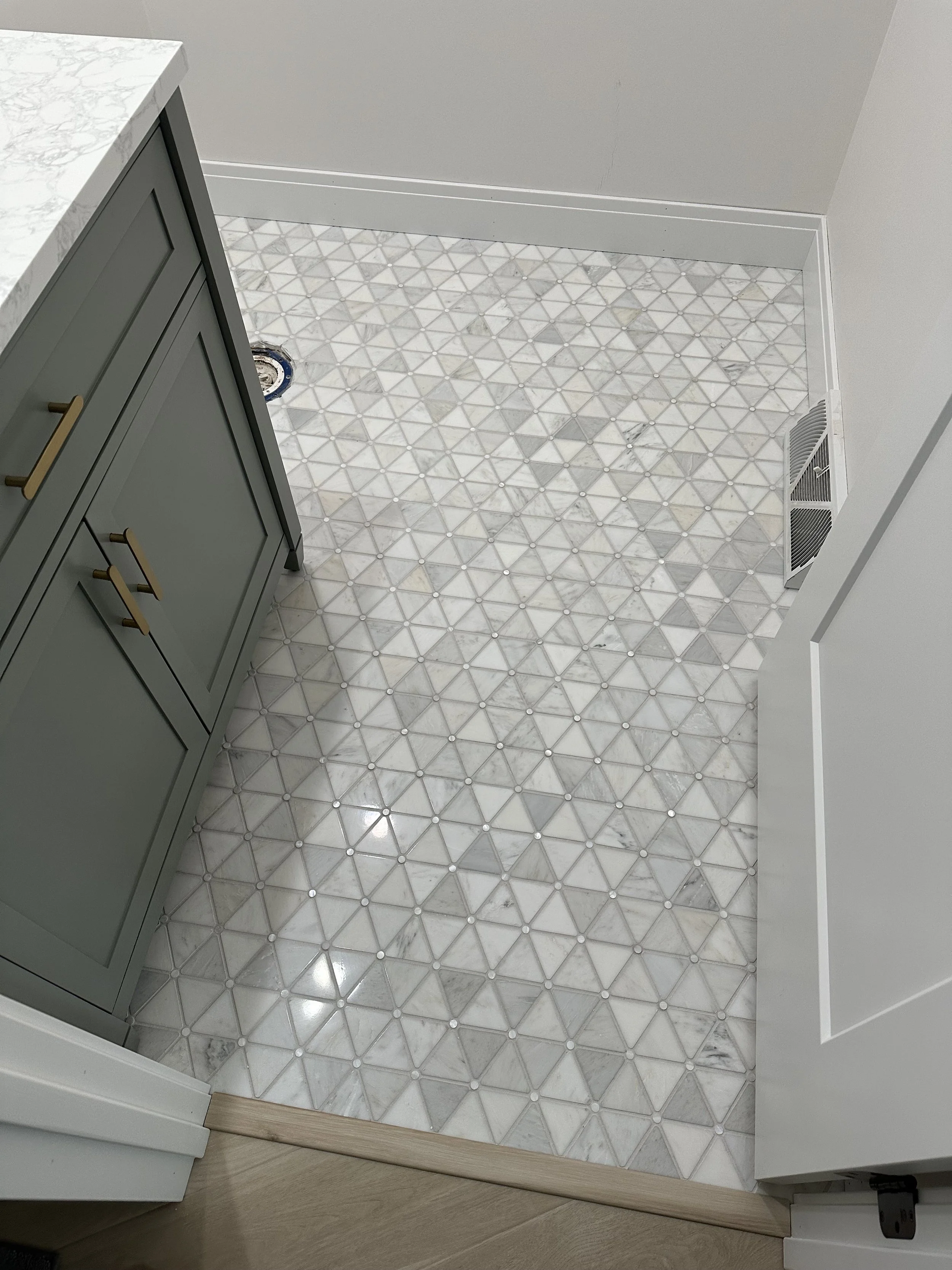 Interior view of a room with patterned white marble floor tiles, a gray cabinet with gold handles, a marble countertop, and a vent on the wall.