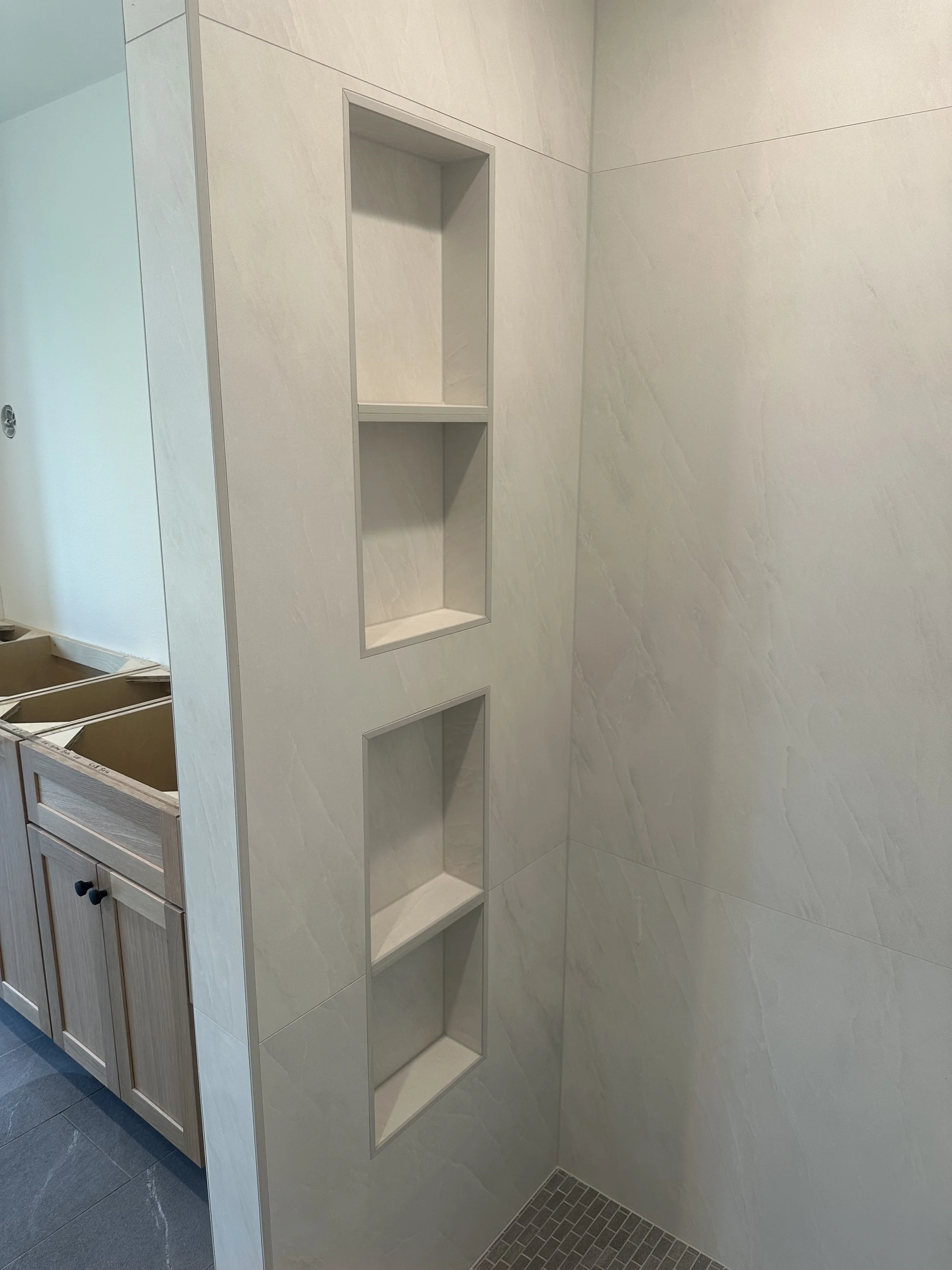 A tiled shower wall with built-in shelves and partial view of a wooden bathroom vanity with cabinets and drawers.