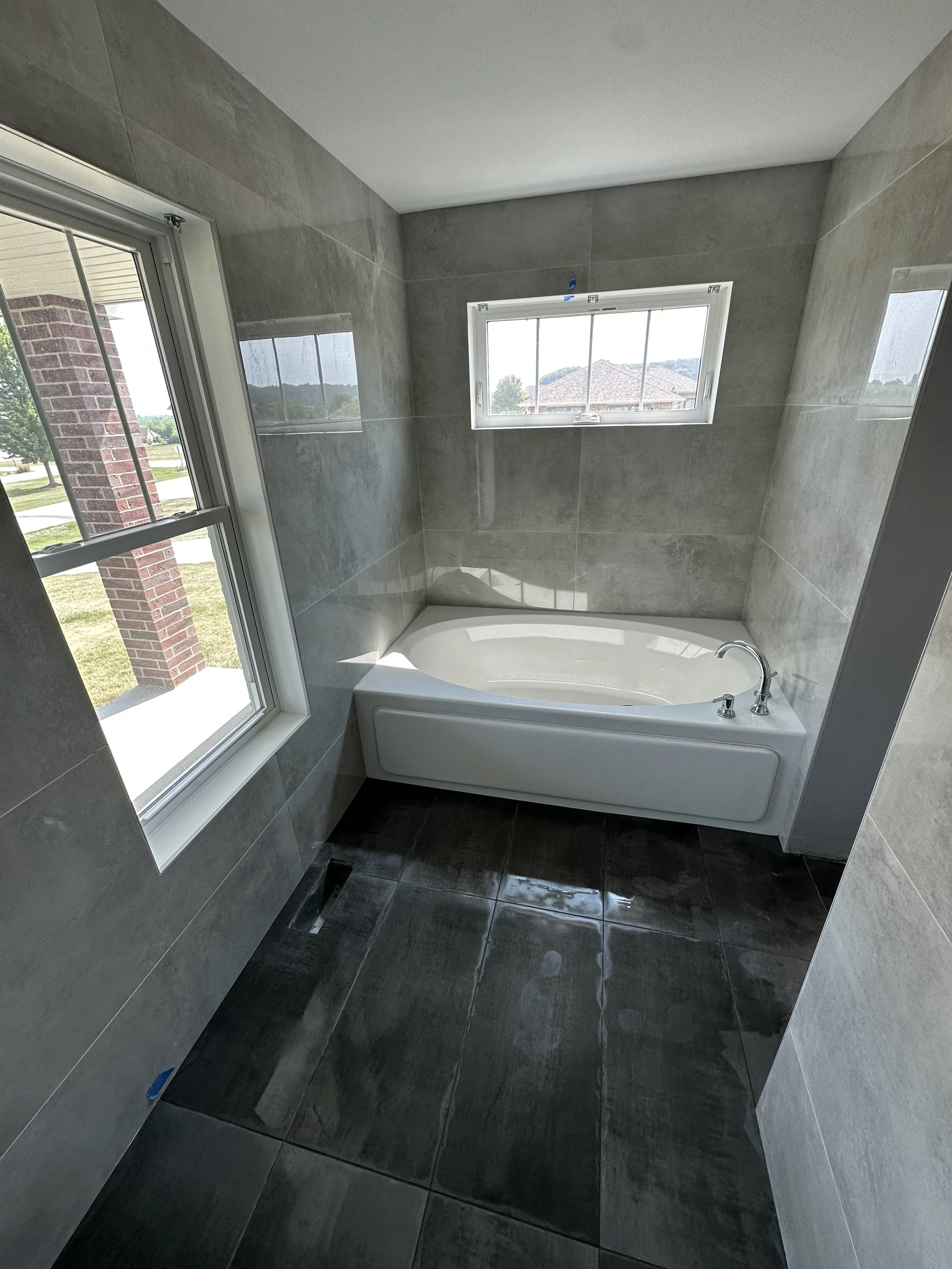 Bathroom with gray tiled walls, black tiled floor, a white bathtub with a silver faucet, and two windows showing a house roof, trees, and a cloudy sky.
