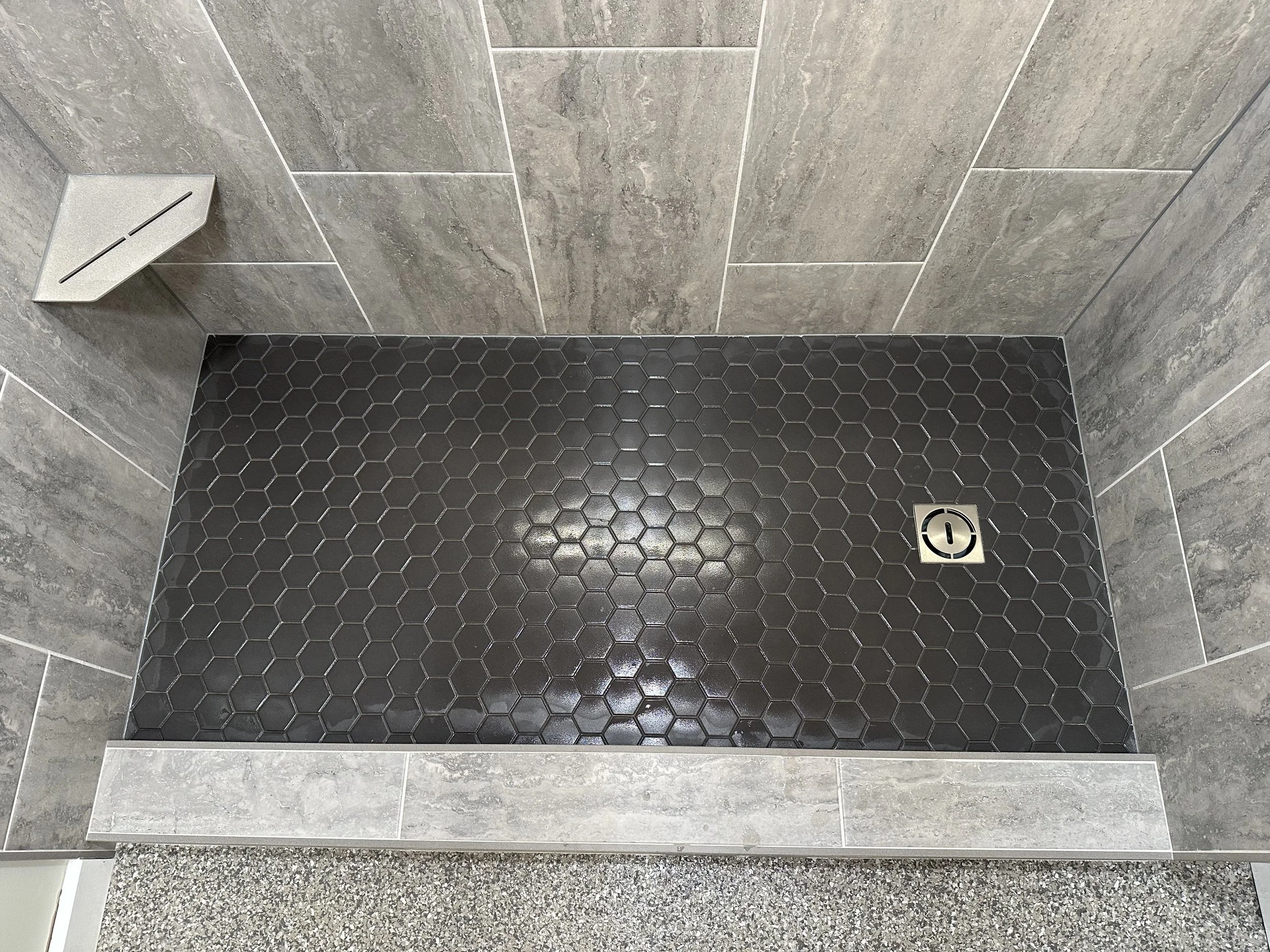 Close-up of a tiled floor with large gray tiles on one side and small black hexagonal tiles on the other, separated by a metal drain.