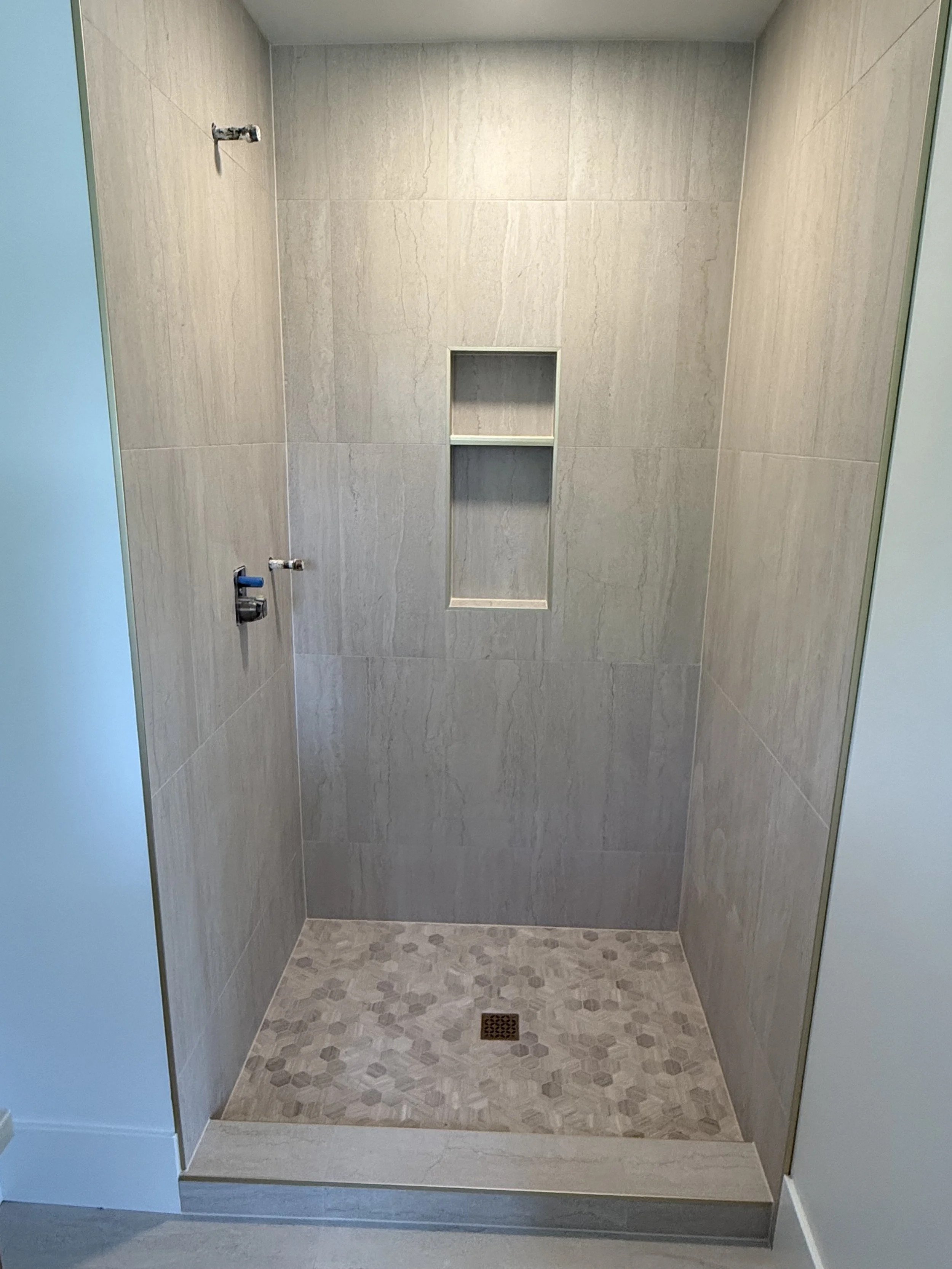 Empty shower with beige tiled walls, small built-in shelf with two sections, and a drain on the patterned floor. No showerhead or fixtures visible.