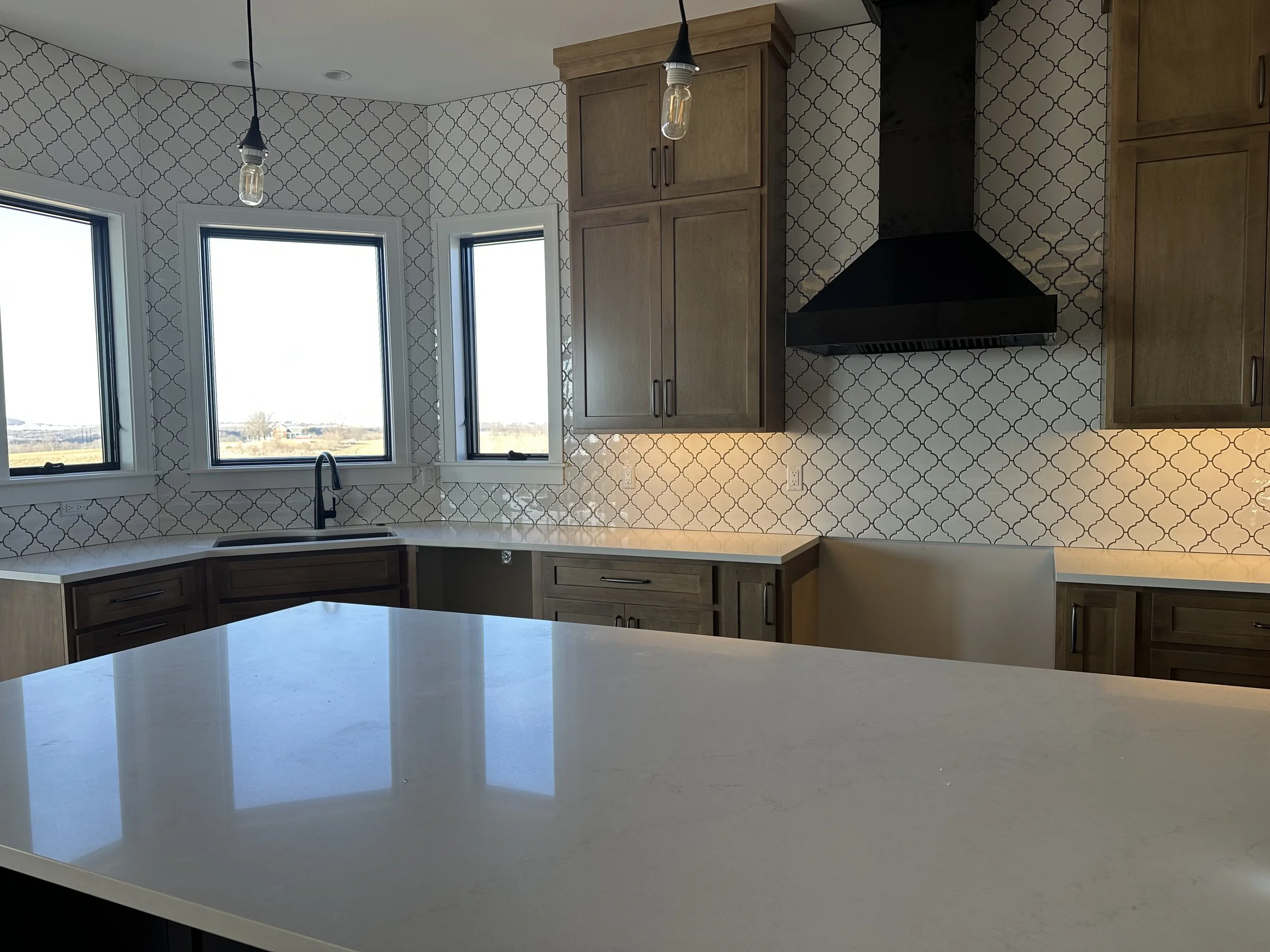 Kitchen with white island countertop, wooden cabinets, black range hood, and patterned backsplash, illuminated by natural light through three windows.