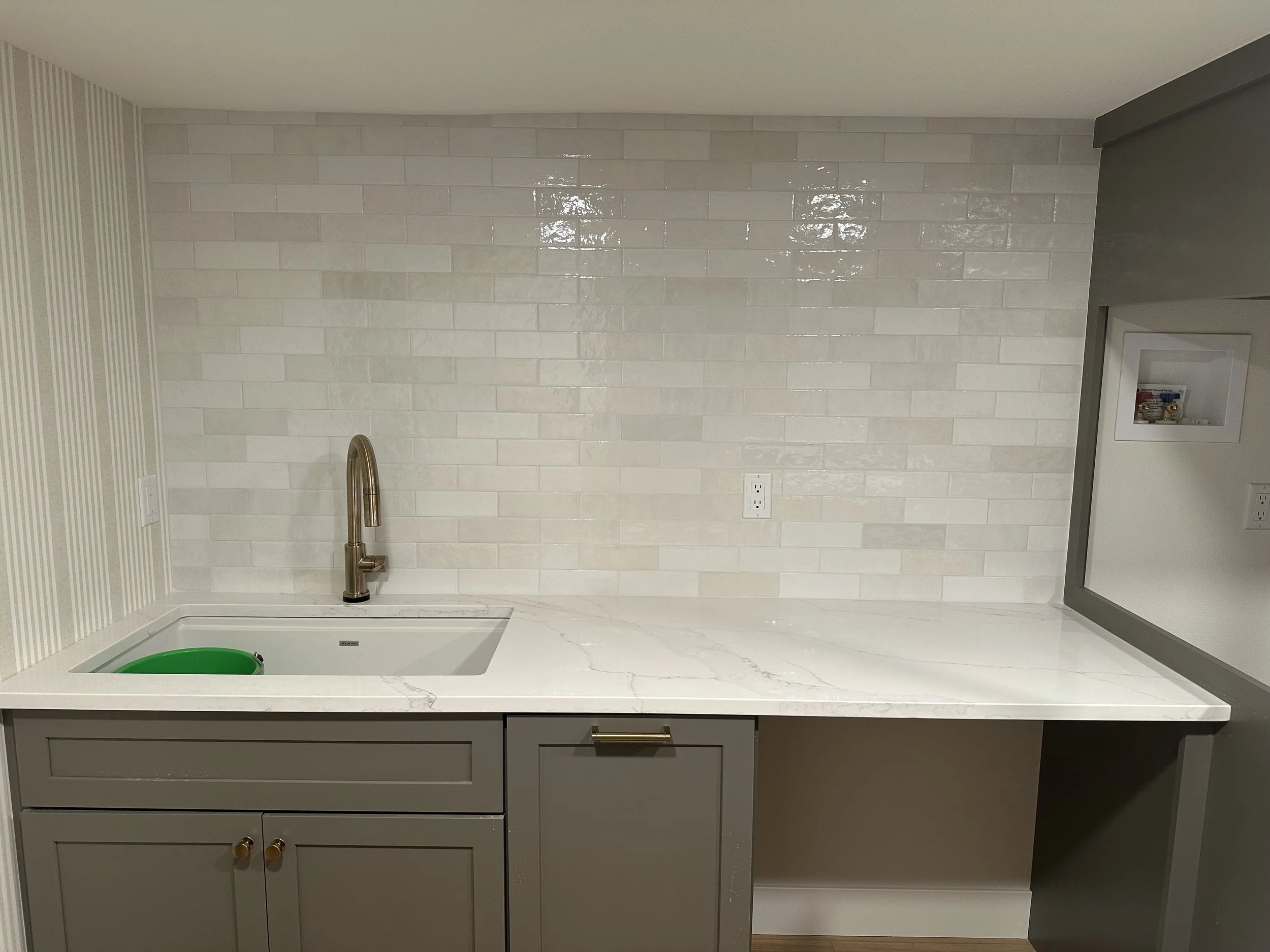 Modern kitchen countertop with gray cabinets, a white marble surface, and a sink with a faucet. The backsplash features glossy beige subway tiles, and there are electrical outlets used for appliances.