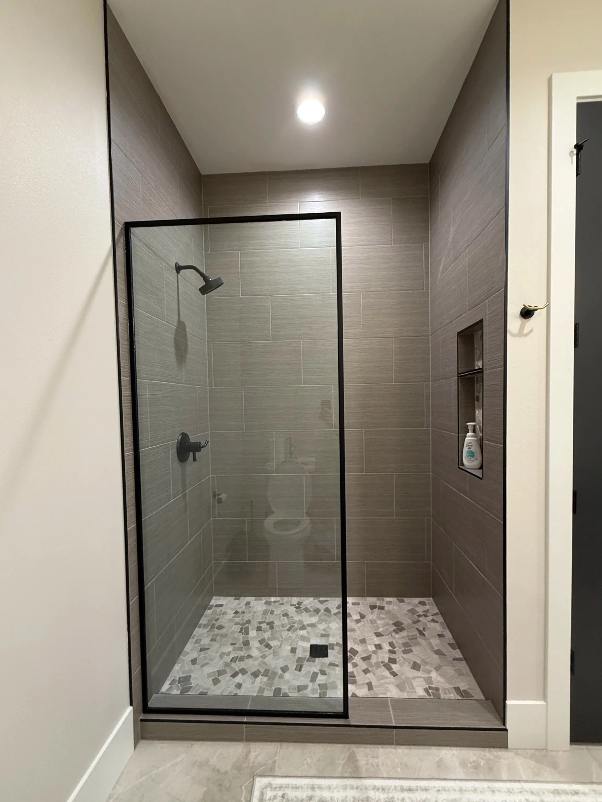 A modern walk-in shower with gray tiles, glass door, and a built-in niche for toiletries, with a white bottle inside. The floor has a mosaic pattern, and a ceiling light illuminates the space.