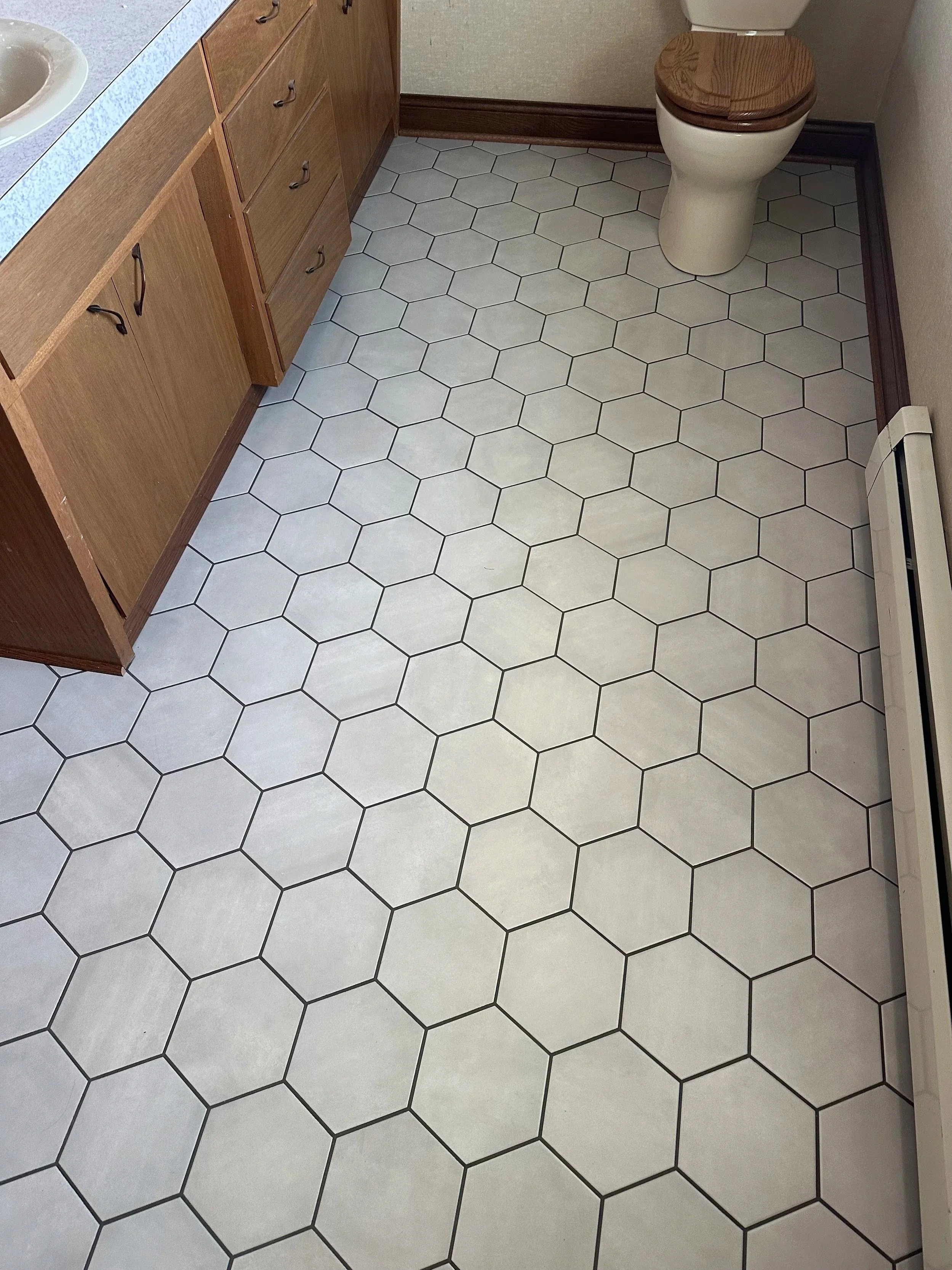 A bathroom with hexagonal white floor tiles outlined in black, wooden cabinets, a toilet with a wooden seat, and a radiator heater along the wall.
