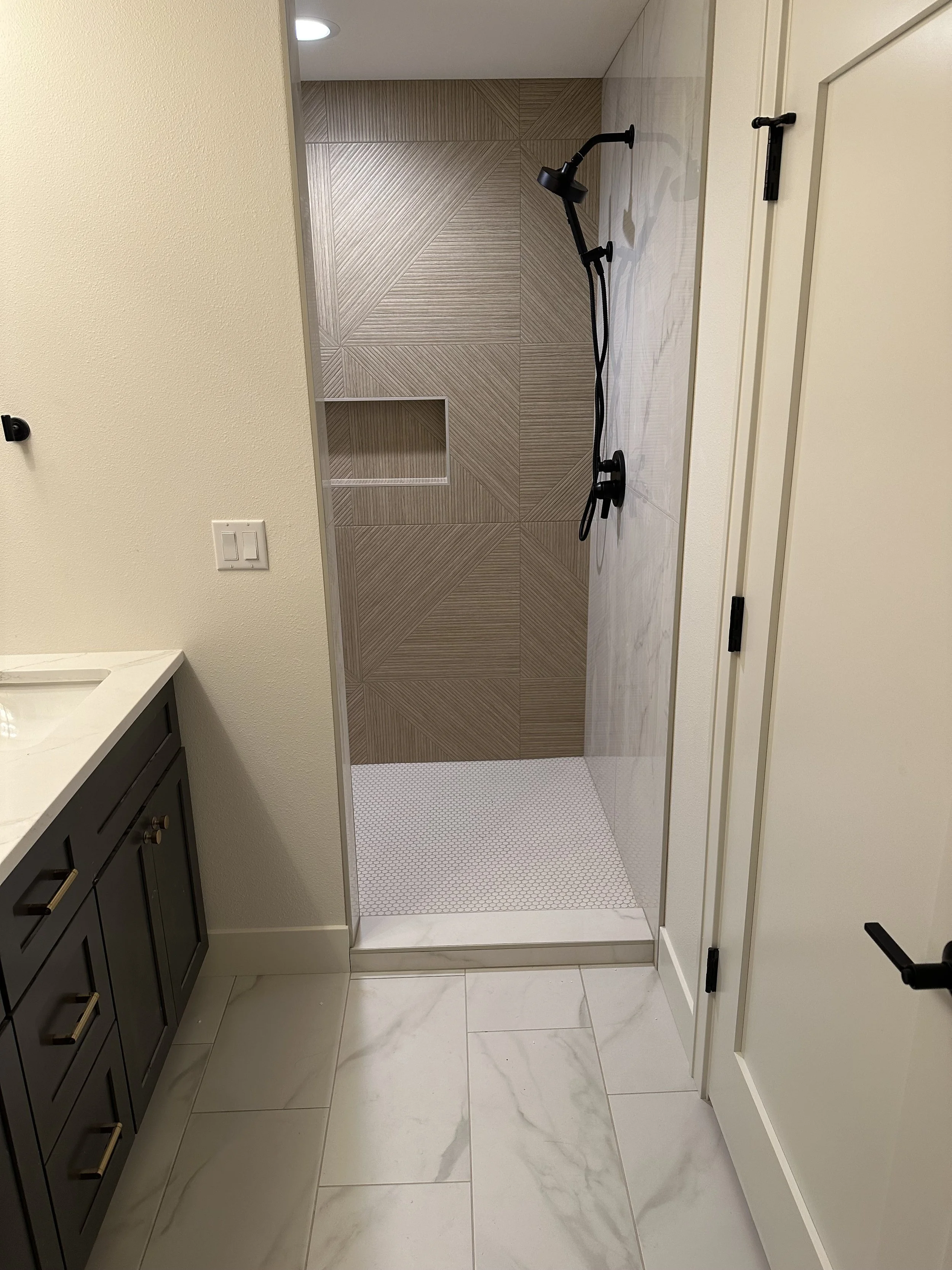 Bathroom with a walk-in shower with patterned wall tiles, a black shower head and controls, a white tiled floor, a dark vanity with gold handles, and beige and white marble floor tiles.