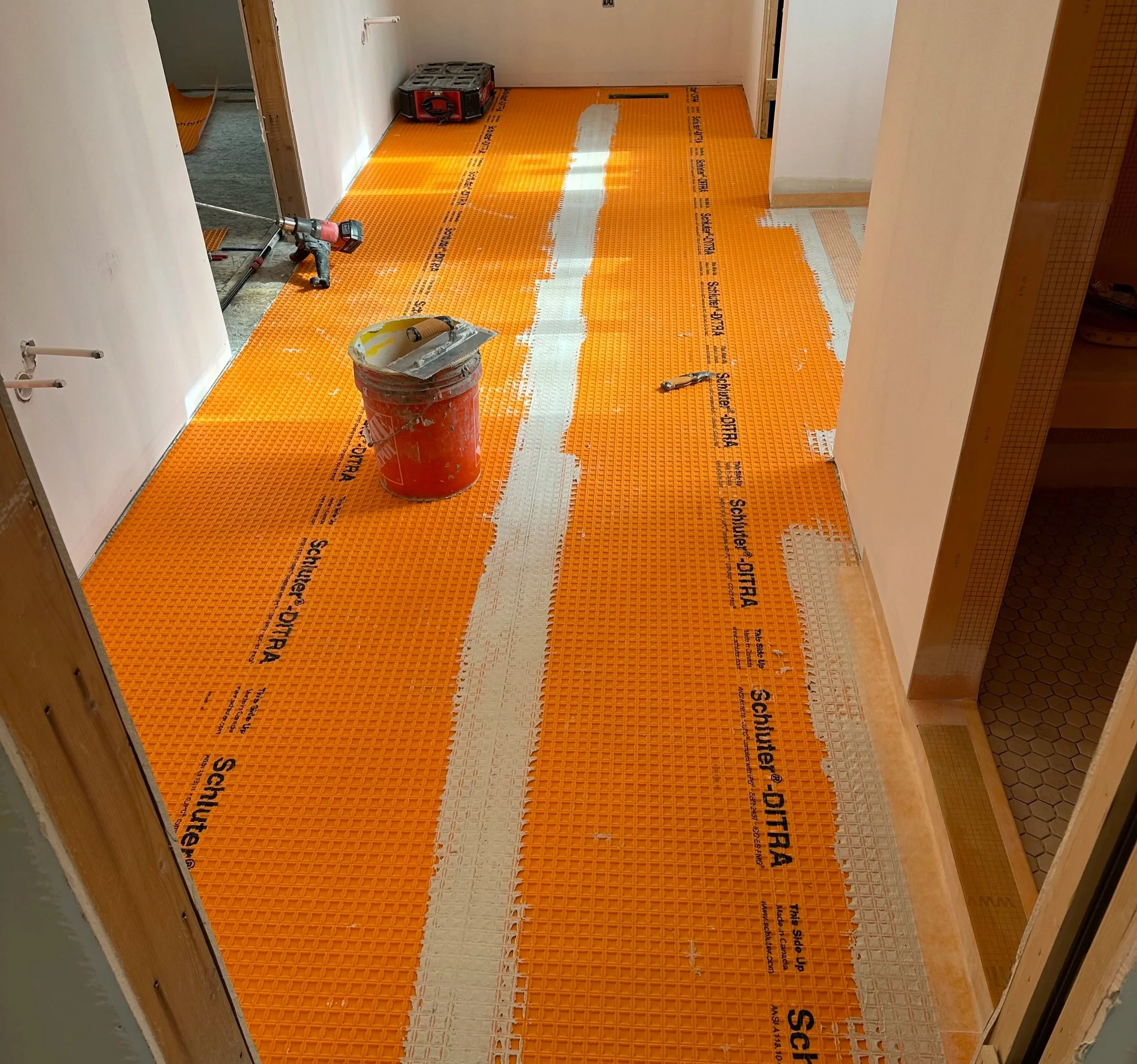 Room with orange plastic flooring for construction, some tools and a bucket on the floor, partially covered walls, and an open doorway.