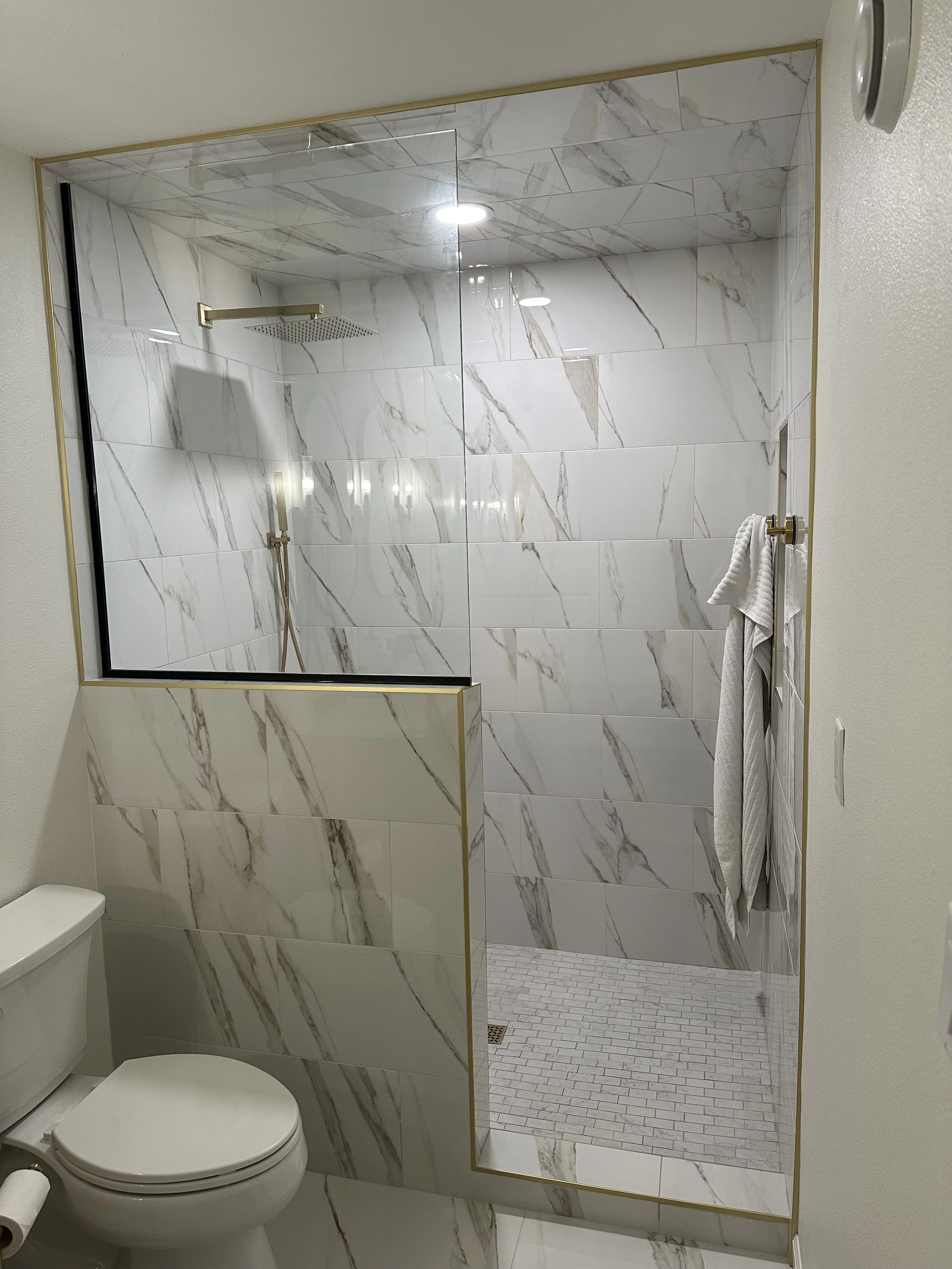 A modern bathroom with a white toilet, a large marble-patterned tile shower with a glass door, and a wall mirror with a gold frame reflecting the shower area.