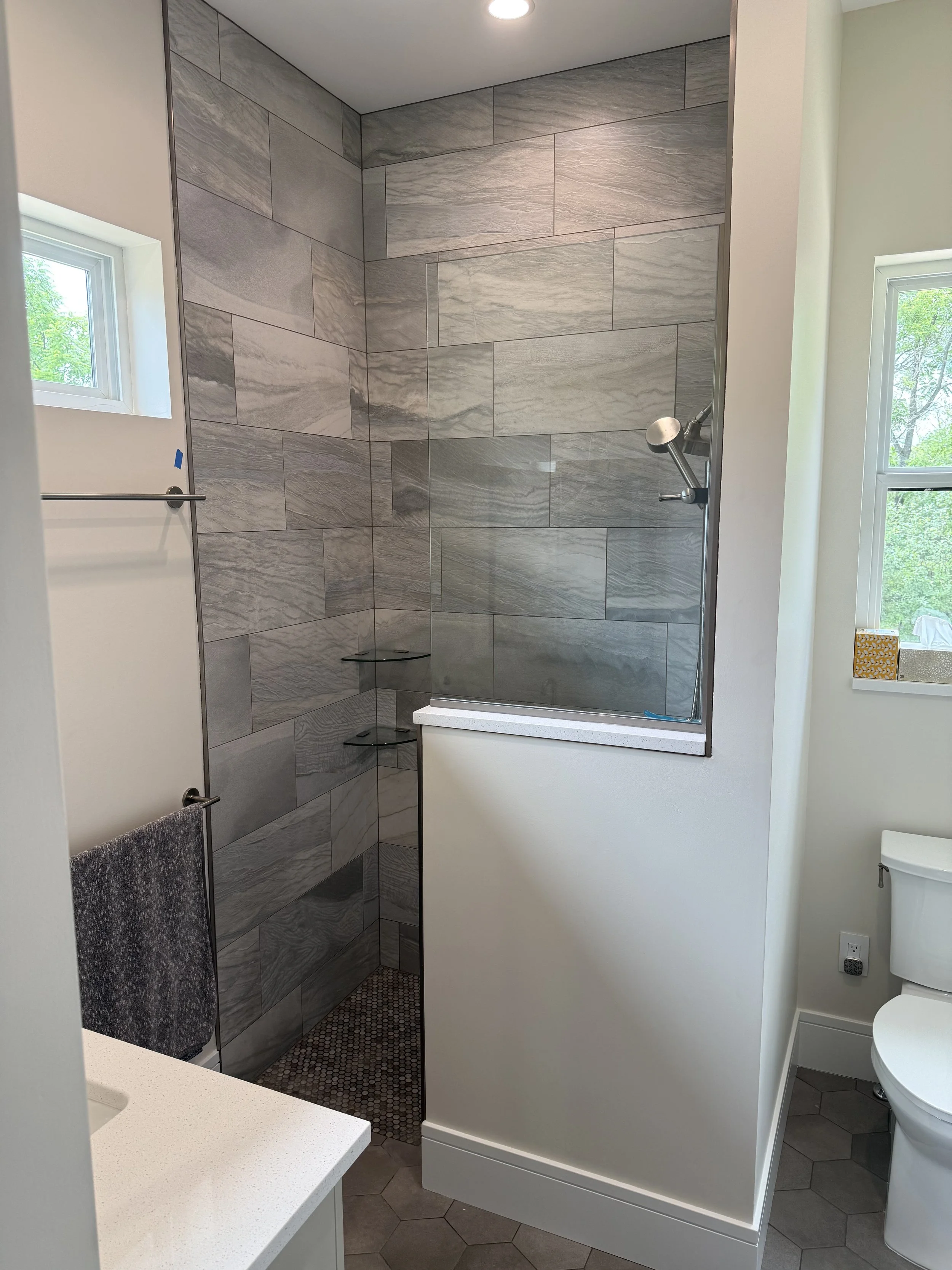 Bathroom shower with gray slate tiles, a small shelf, and a hand-held showerhead. Next to the shower is a white wall with two windows, a towel hanging, and a bathroom toilet.