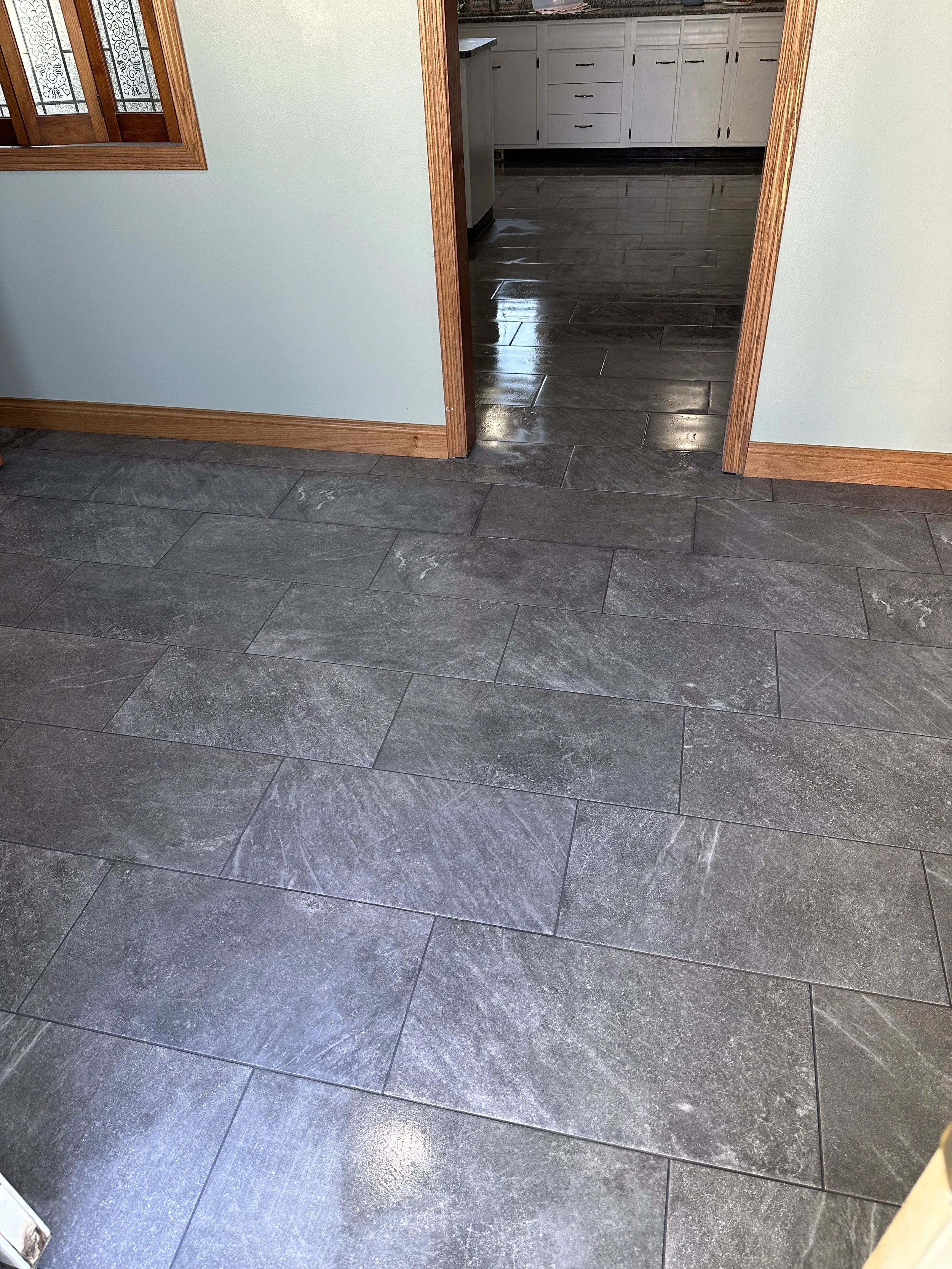 A kitchen with dark gray tiled flooring, white cabinets with black handles, and a counter with a granite surface. An open doorway leads to the kitchen area.