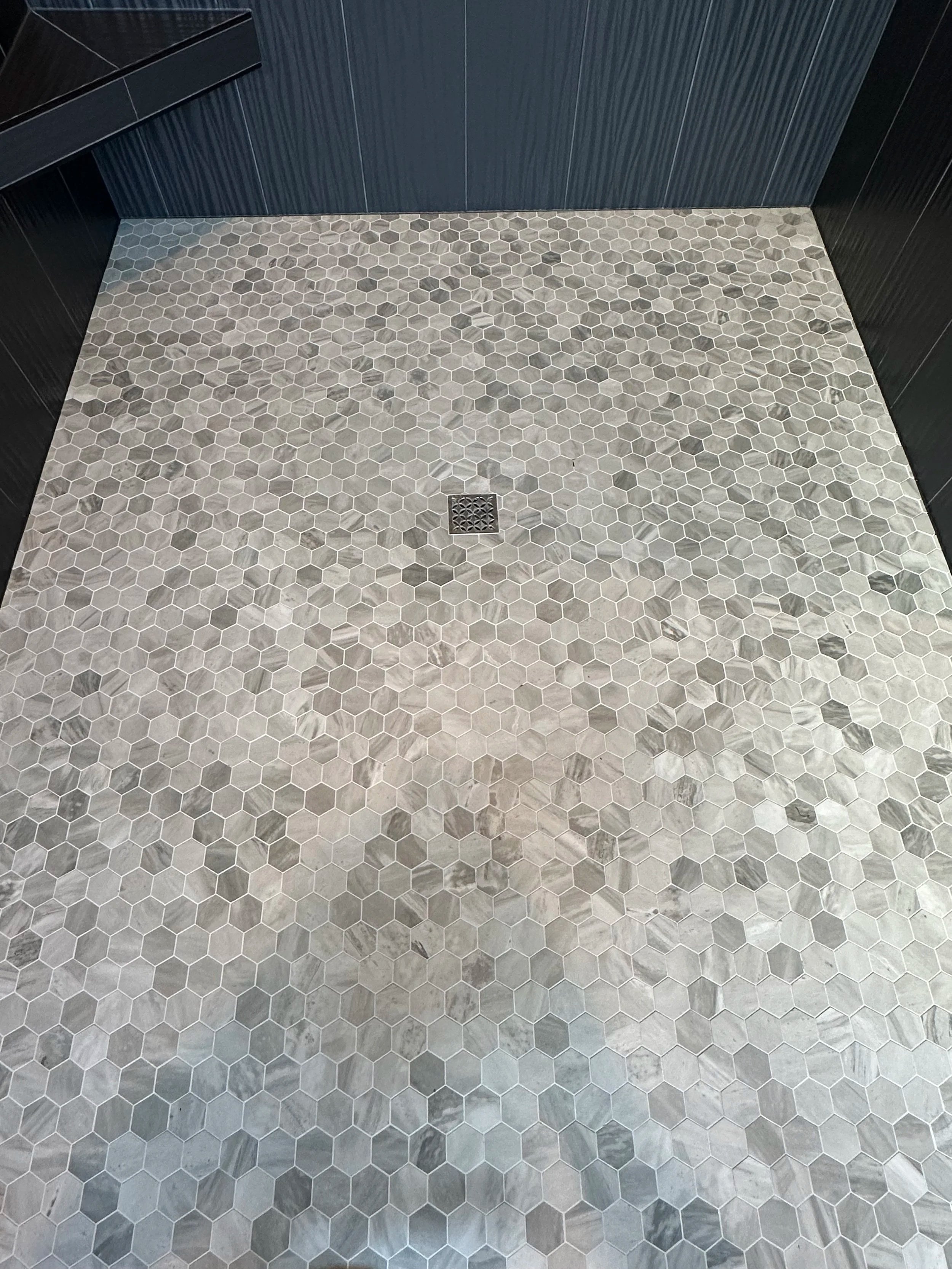 A bathroom floor with hexagonal gray tiles and a central metal drain grate, surrounded by black walls.