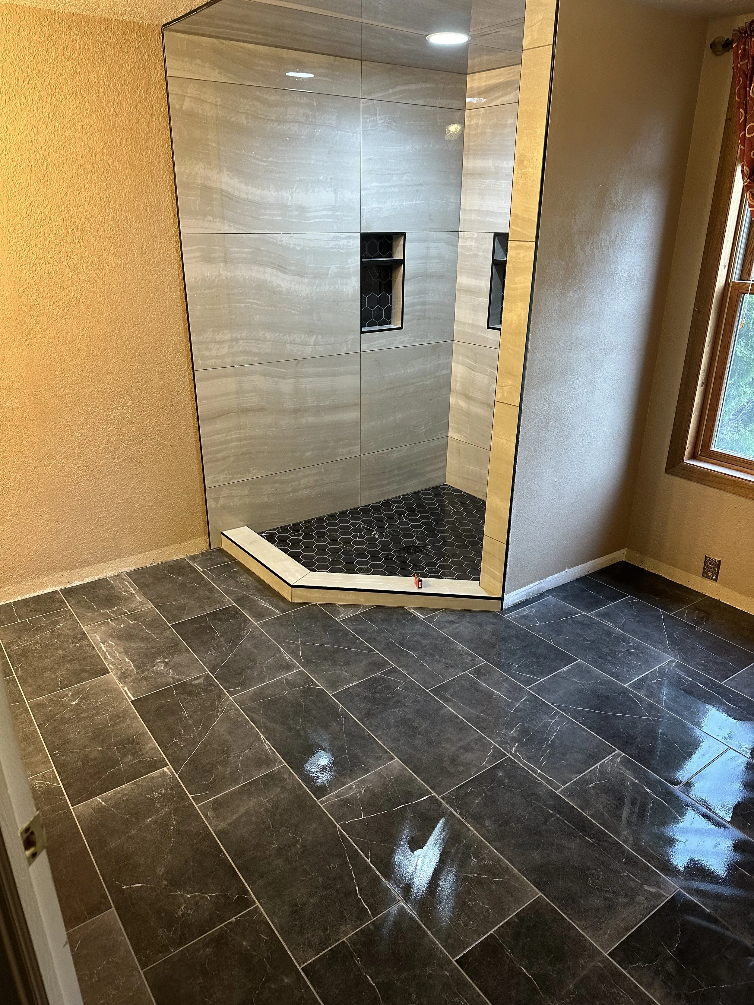 A bathroom shower area with gray tiled walls and black hexagonal floor tiles, illuminated by recessed ceiling lights and adjacent to a window with a wood frame.
