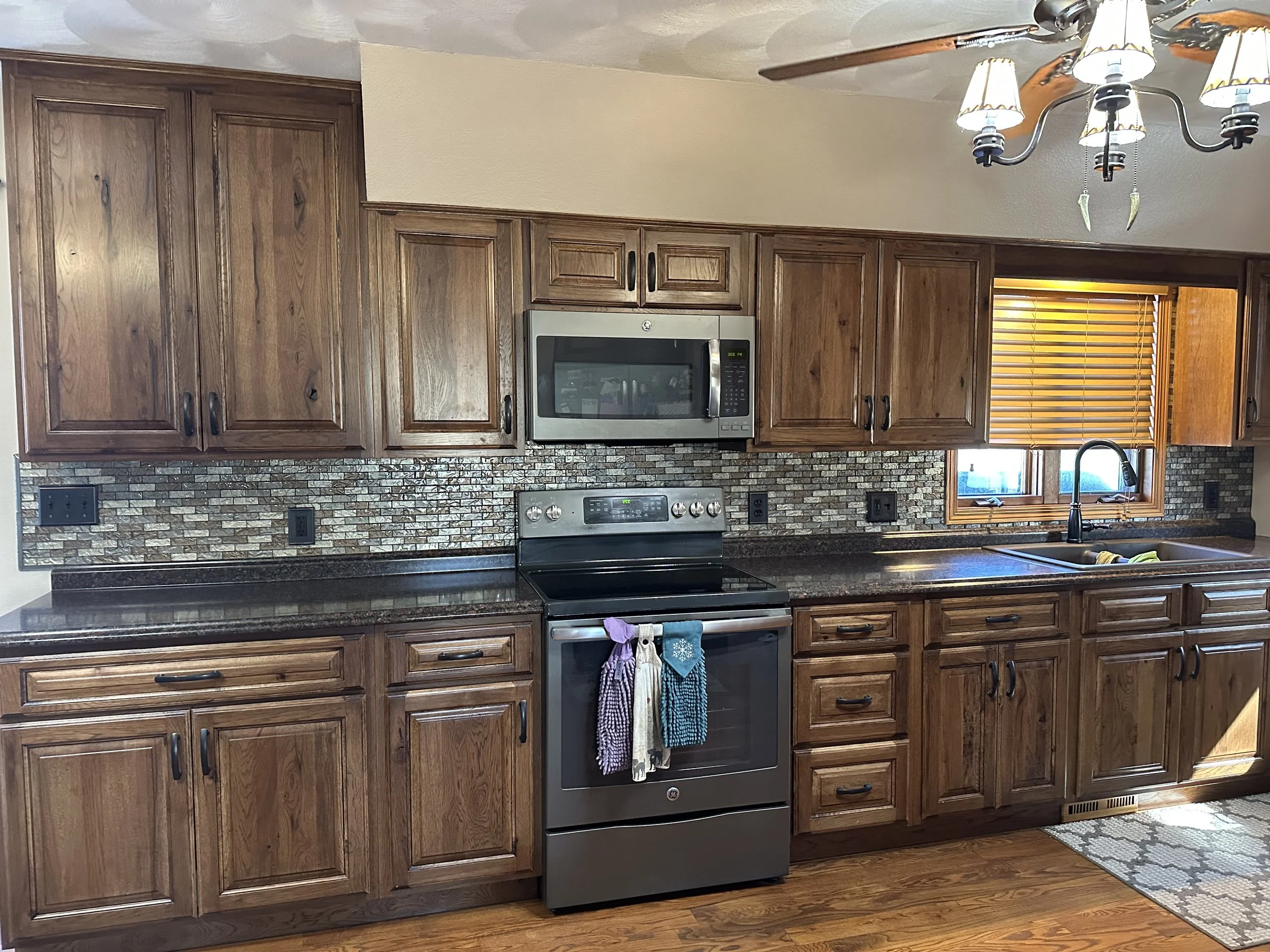 Kitchen with wooden cabinets, stainless steel microwave and oven, brick backsplash, and a window with wooden blinds. There are kitchen towels hanging in front of the oven.