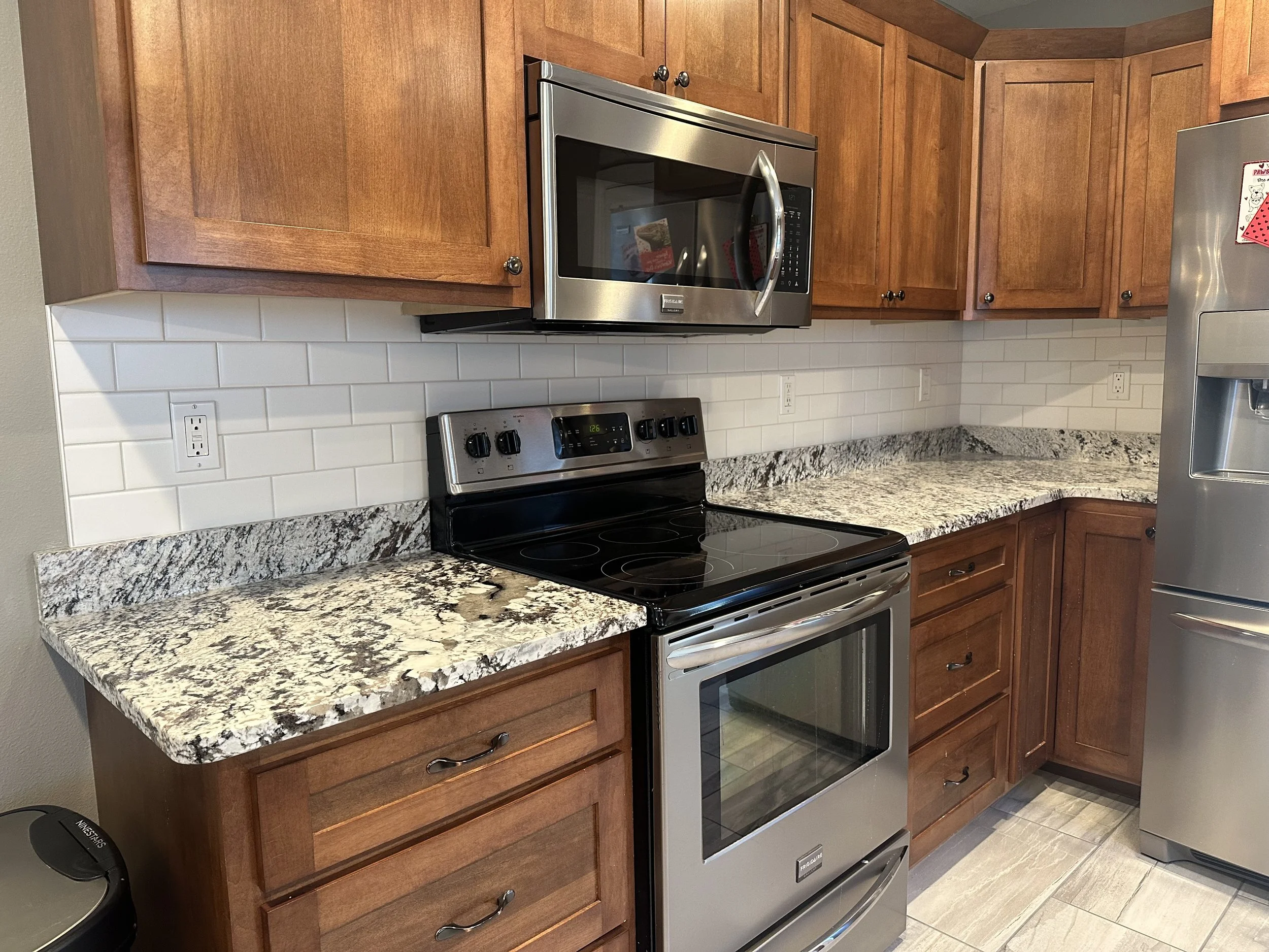 Kitchen with oak cabinets, granite countertops, stainless steel microwave and stove, white Subway tile backsplash, and a partial view of a stainless steel refrigerator.