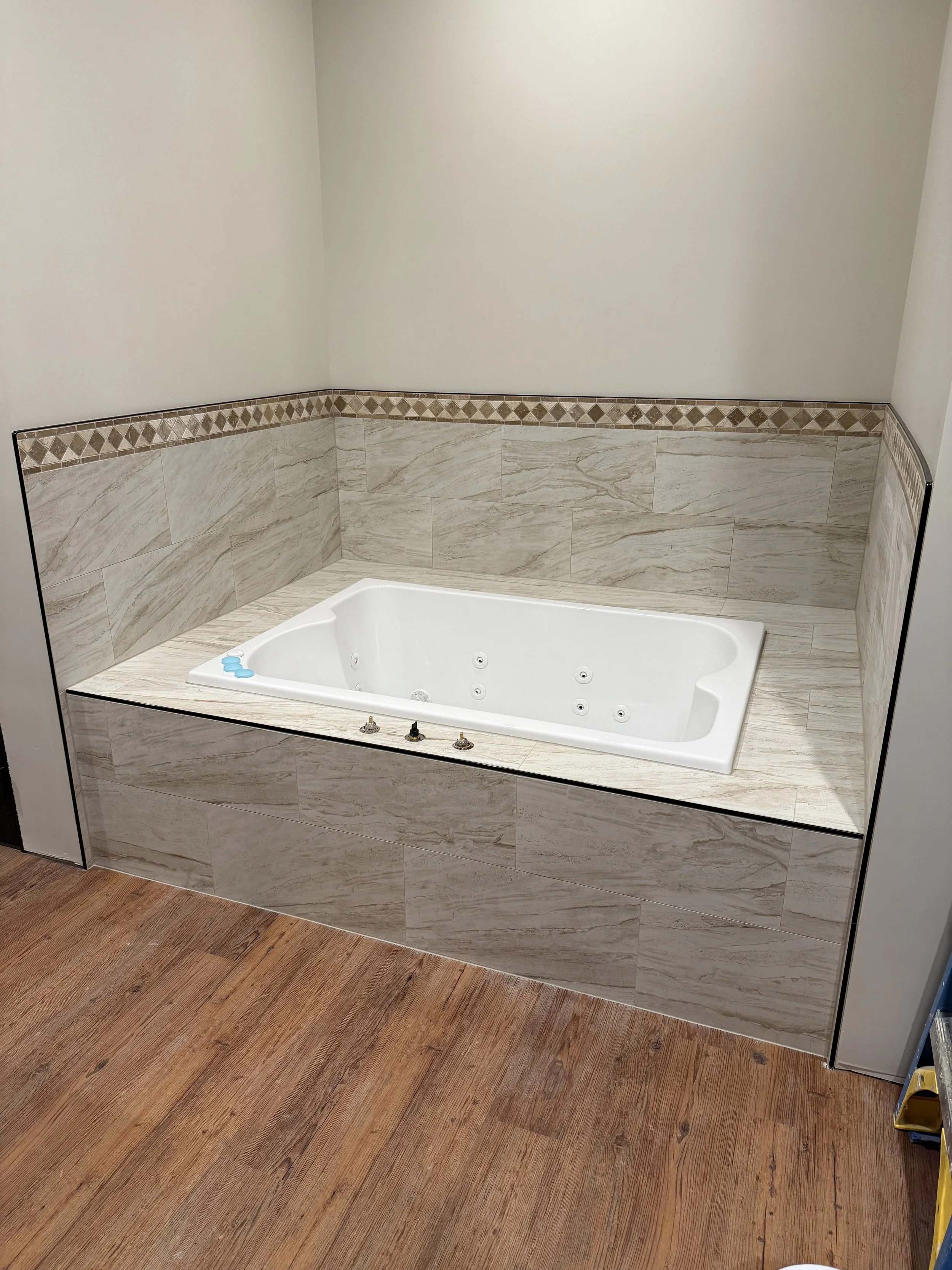 An empty bathroom with a corner whirlpool bathtub, beige marble tile walls with a diamond border, and a wood floor.