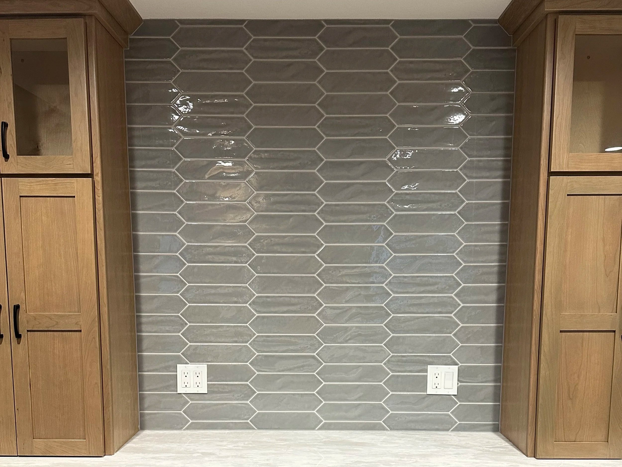 Kitchen backsplash with hexagonal gray tiles, wooden cabinets on each side, and electrical outlets at the bottom.