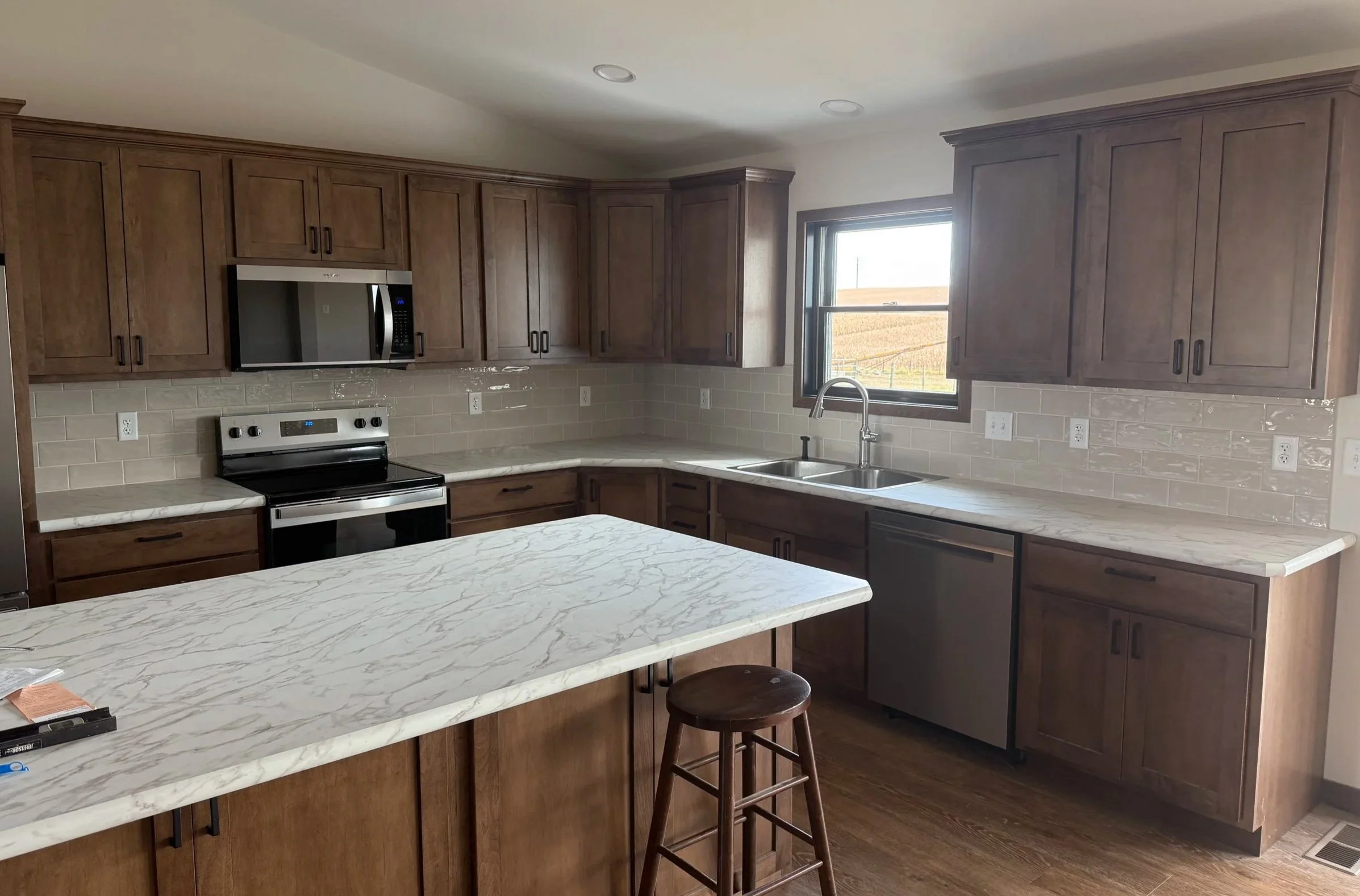 Kitchen with wooden cabinets, marble countertops, a stainless steel microwave and oven, a window with a view of fields, and a kitchen island with a stool.