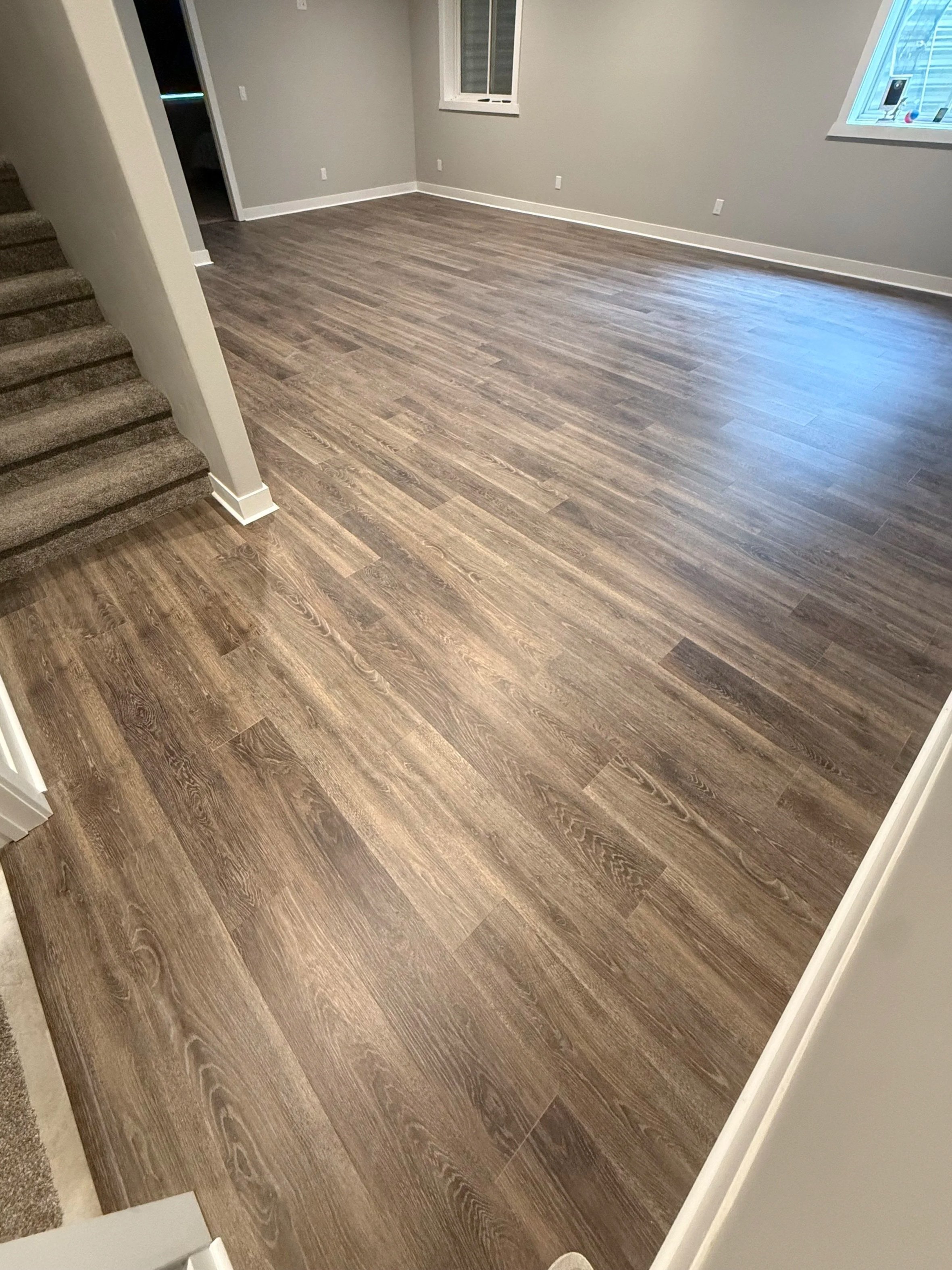 Empty living room with gray walls, white trim, and hardwood floors. There are two windows, one open, and a staircase on the left side.