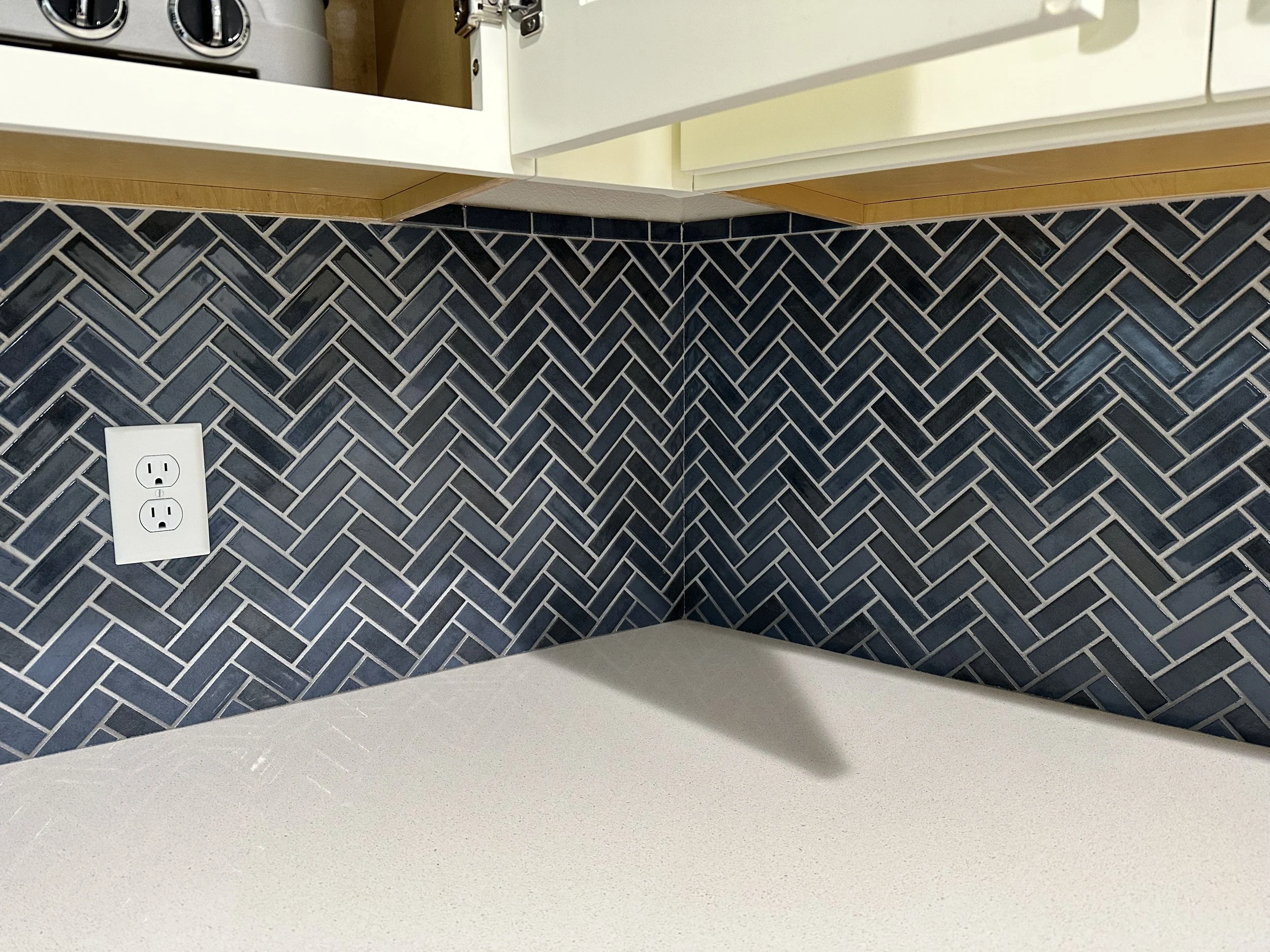 Close-up of a kitchen counter with a black herringbone tile backsplash, a white electrical outlet, and part of a white stove and upper cabinets.
