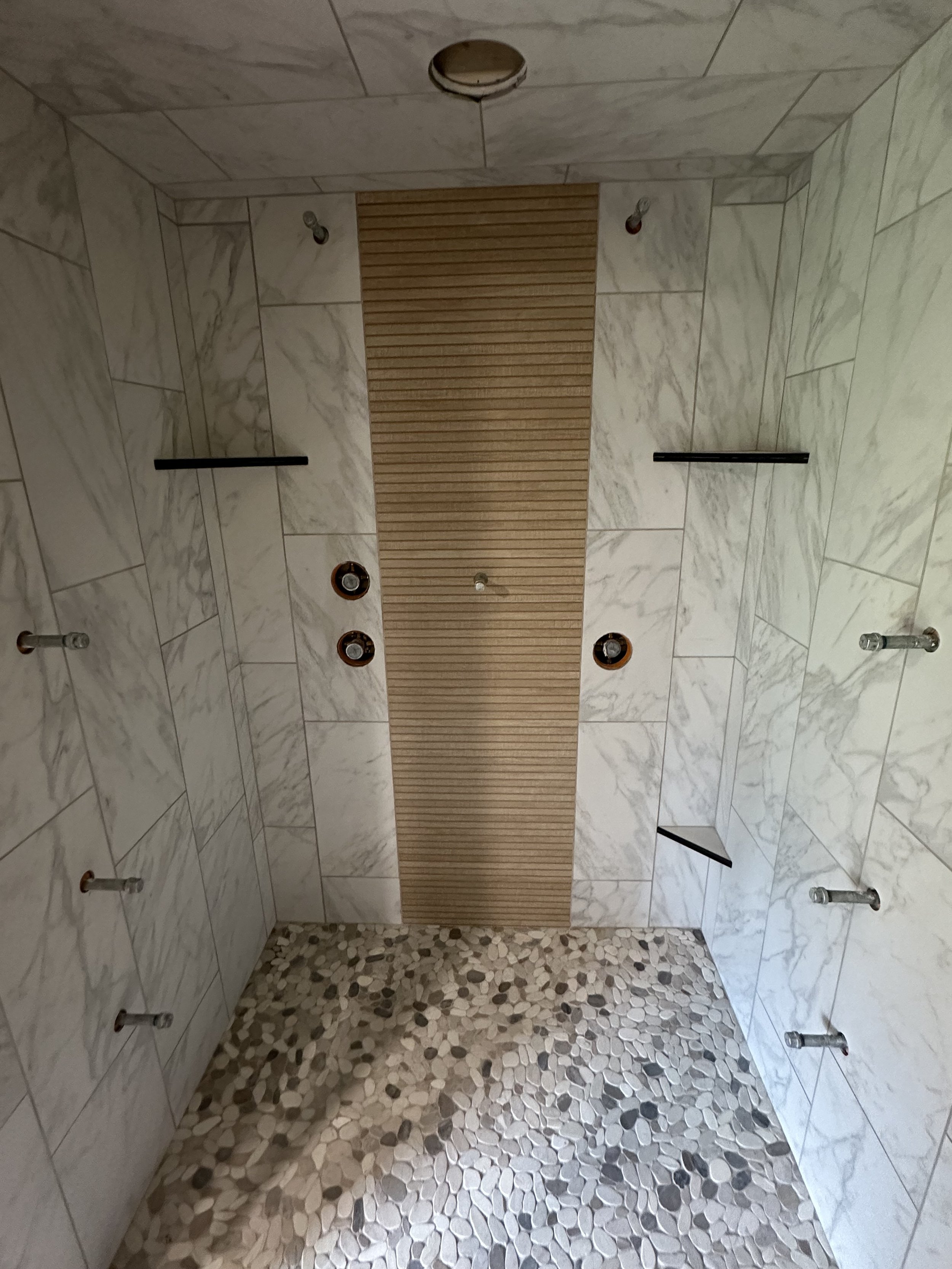 Shower area with white marble tiles, a wooden slat panel on the ceiling, multiple showerheads, and a pebble stone floor.