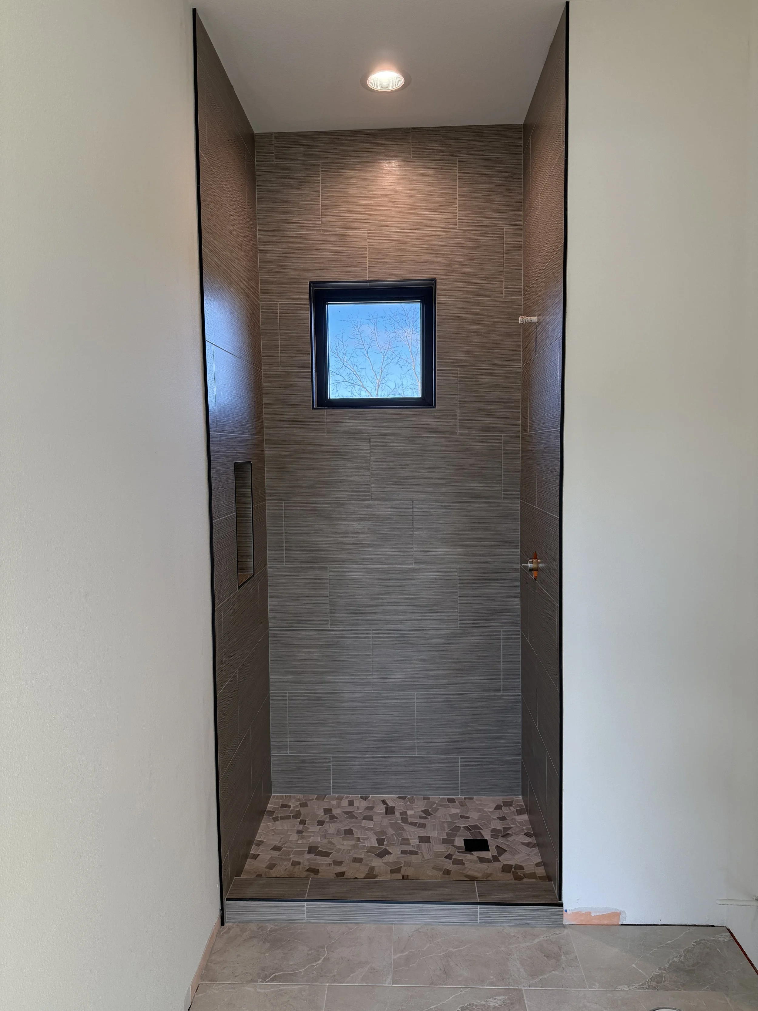 Inside view of a modern shower with brown textured wall tiles, a small window showing blue sky and tree branches, and a tiled floor in small mosaic pattern.