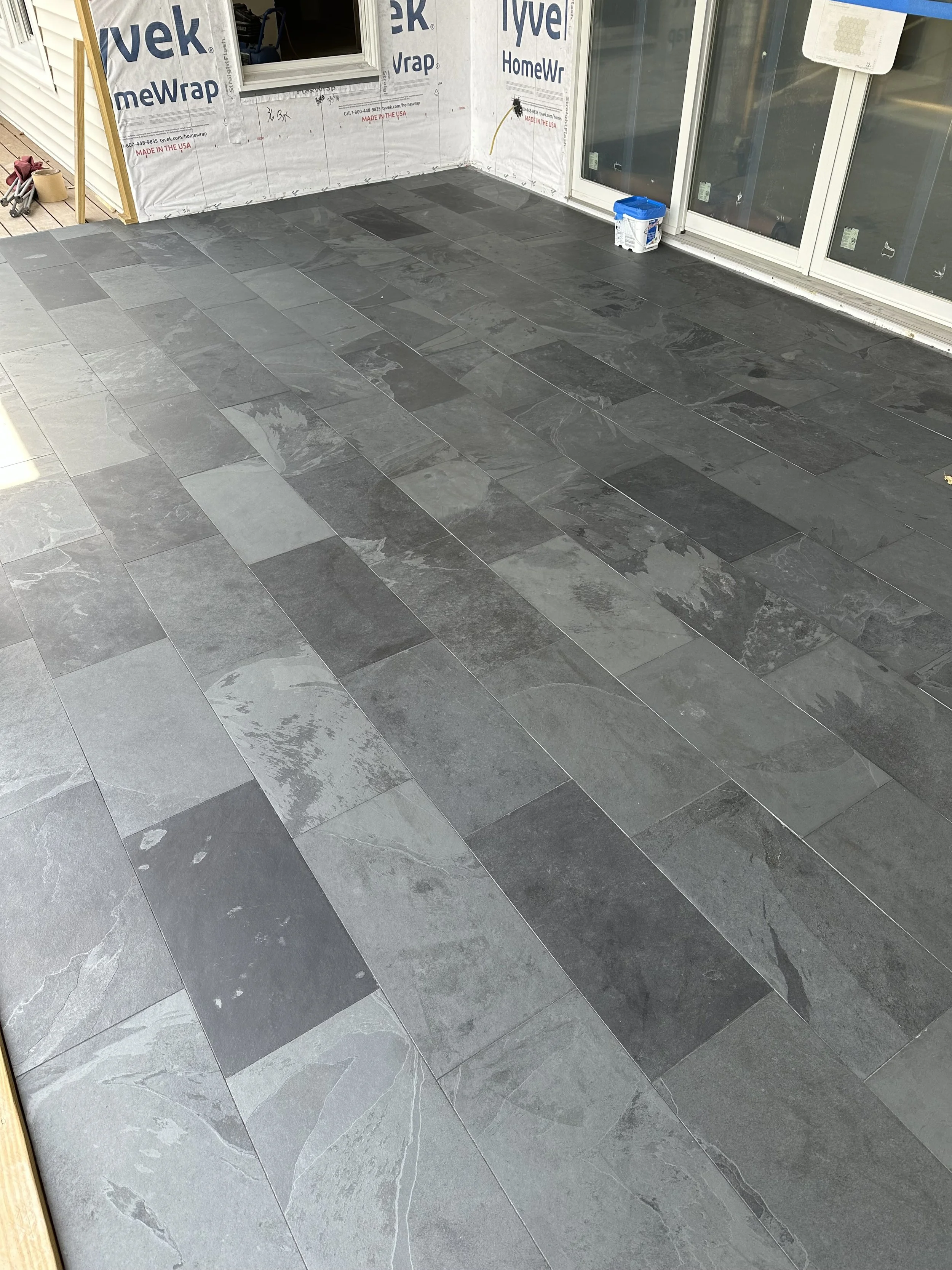 A newly tiled outdoor patio area with dark gray and black stone tiles, adjacent to a house under construction. The house has partially installed siding, a window, a door, and construction materials including a small blue container of paint or sealant on the floor.