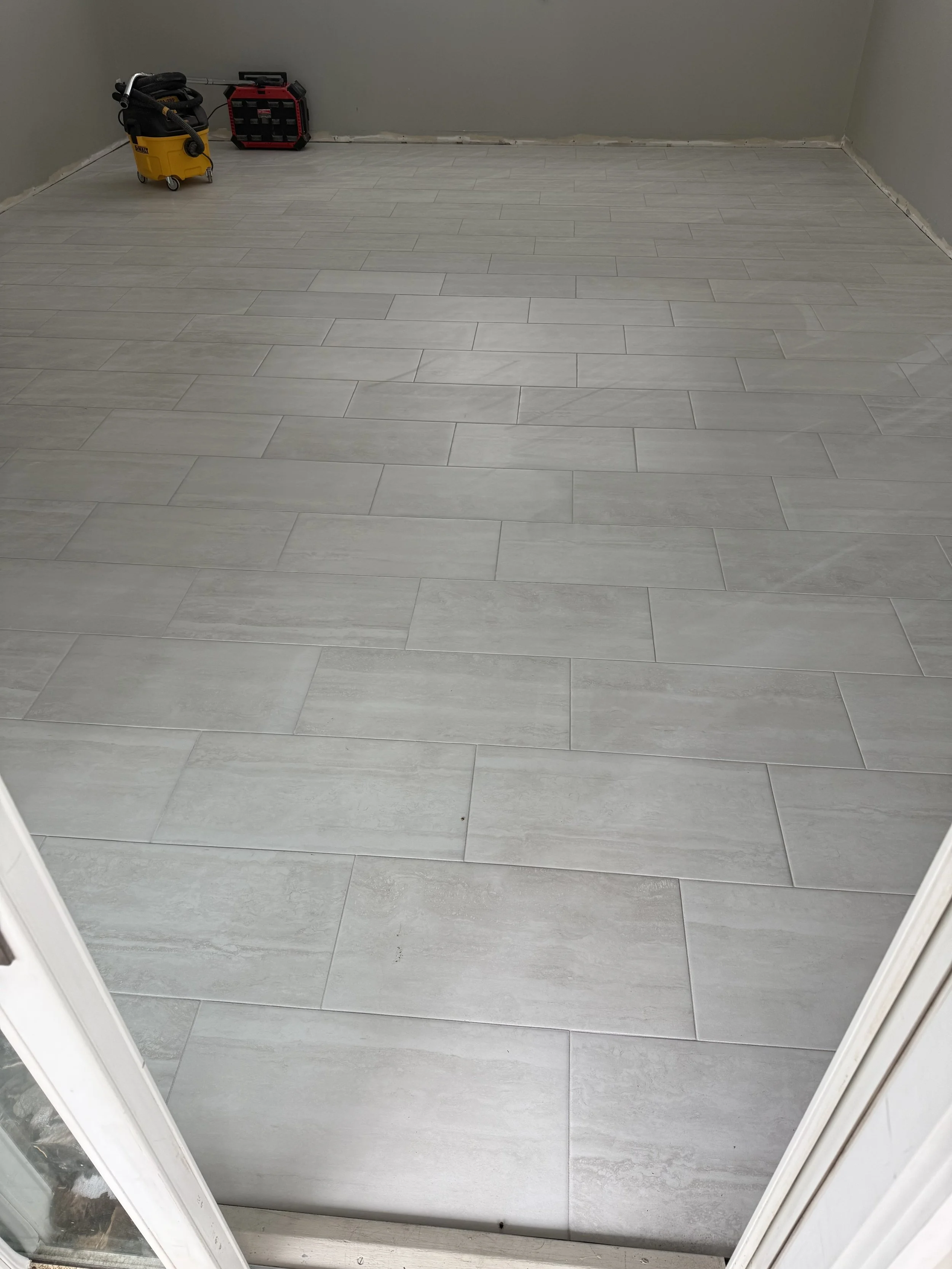 View through a doorway showing a room with newly installed gray tile flooring and unfinished light gray walls. There are construction tools, including a yellow wet/dry vacuum and a black toolbox with a red frame, positioned in the corner of the room.
