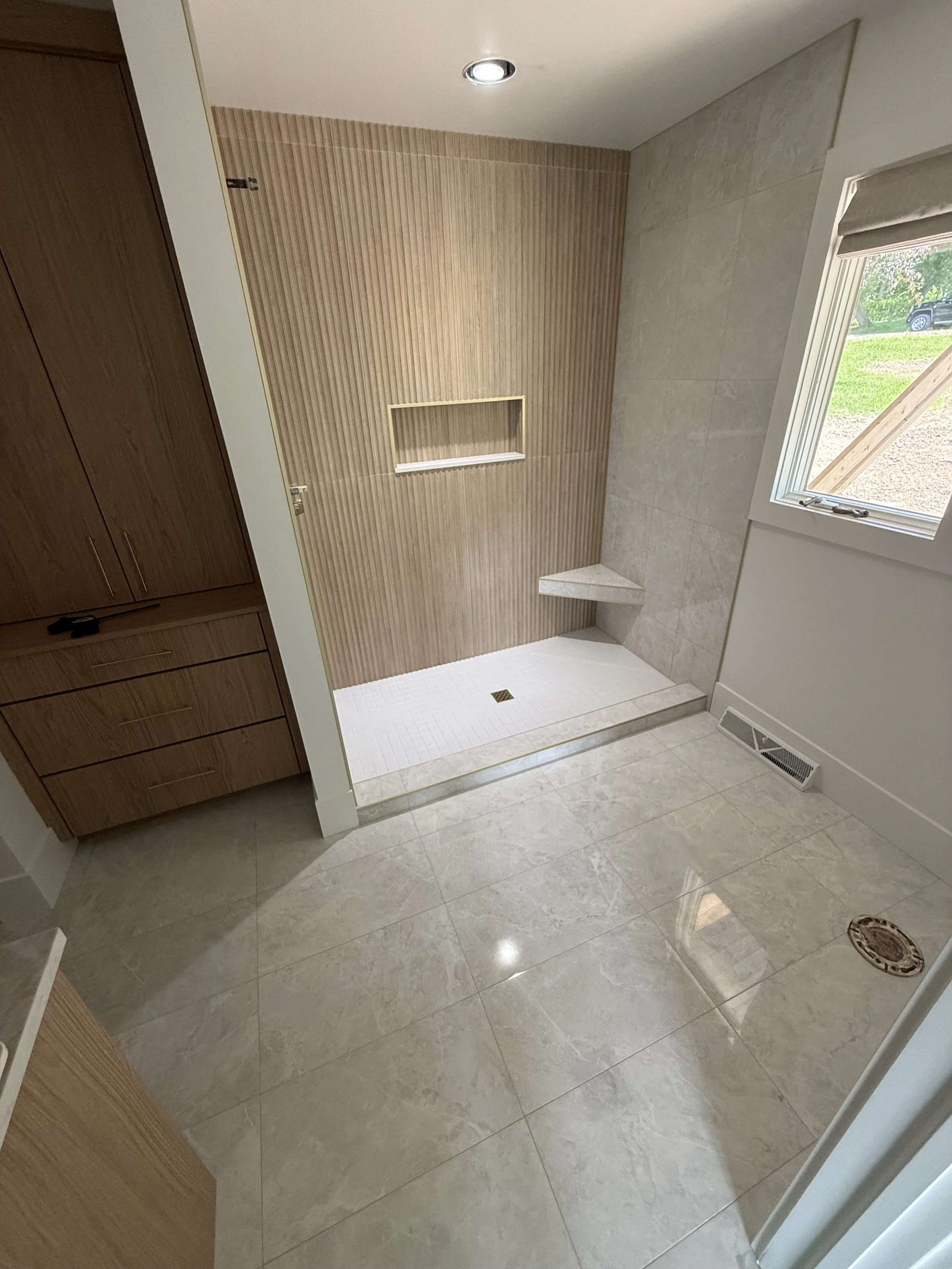 A bathroom with beige tiled floor, a walk-in shower with a partial glass door, a wooden cabinet, and a window showing outdoor greenery.