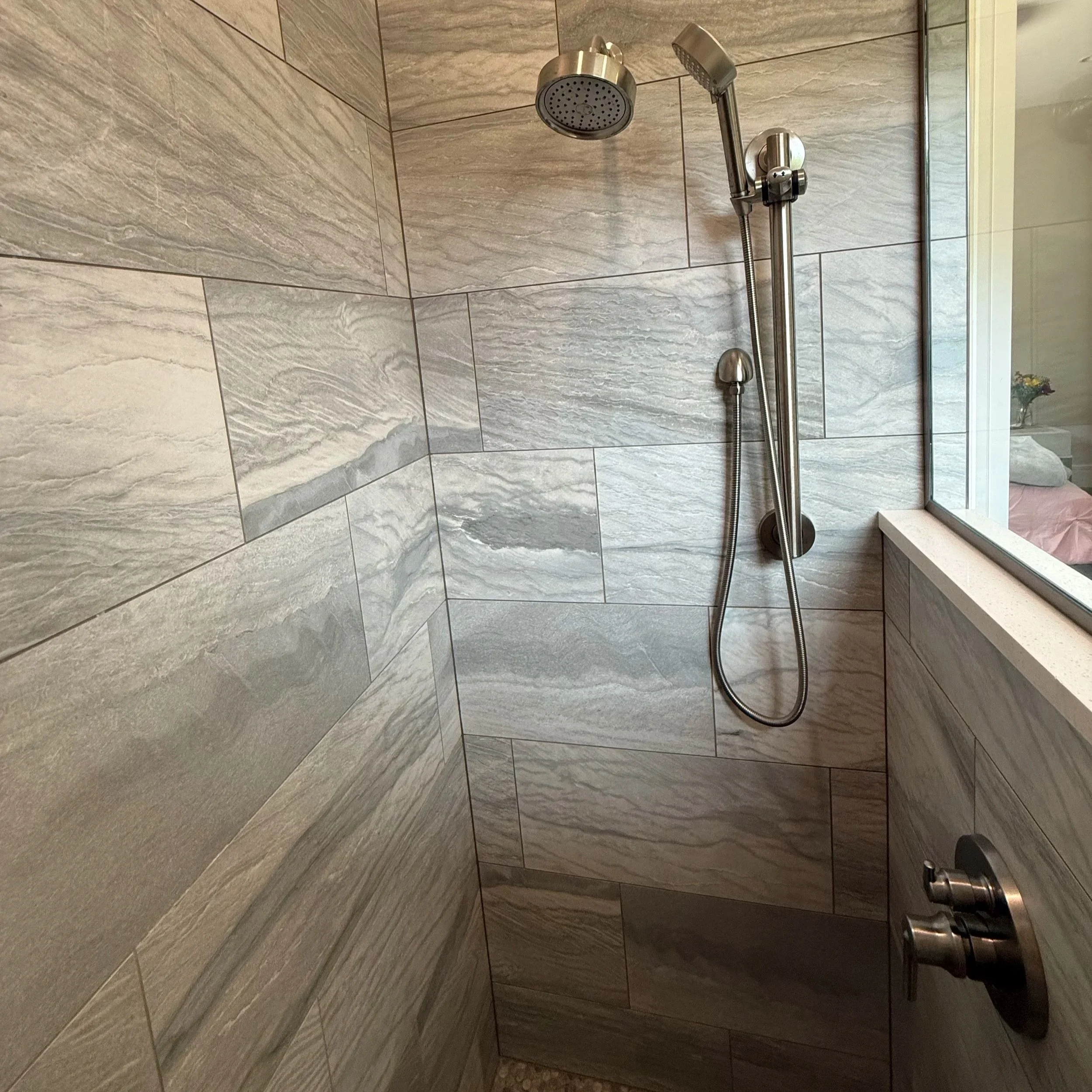 Shower area with large beige and gray stone tiles, a ceiling-mounted showerhead, a handheld shower with a hose, and a large window with a flowerpot on the sill.