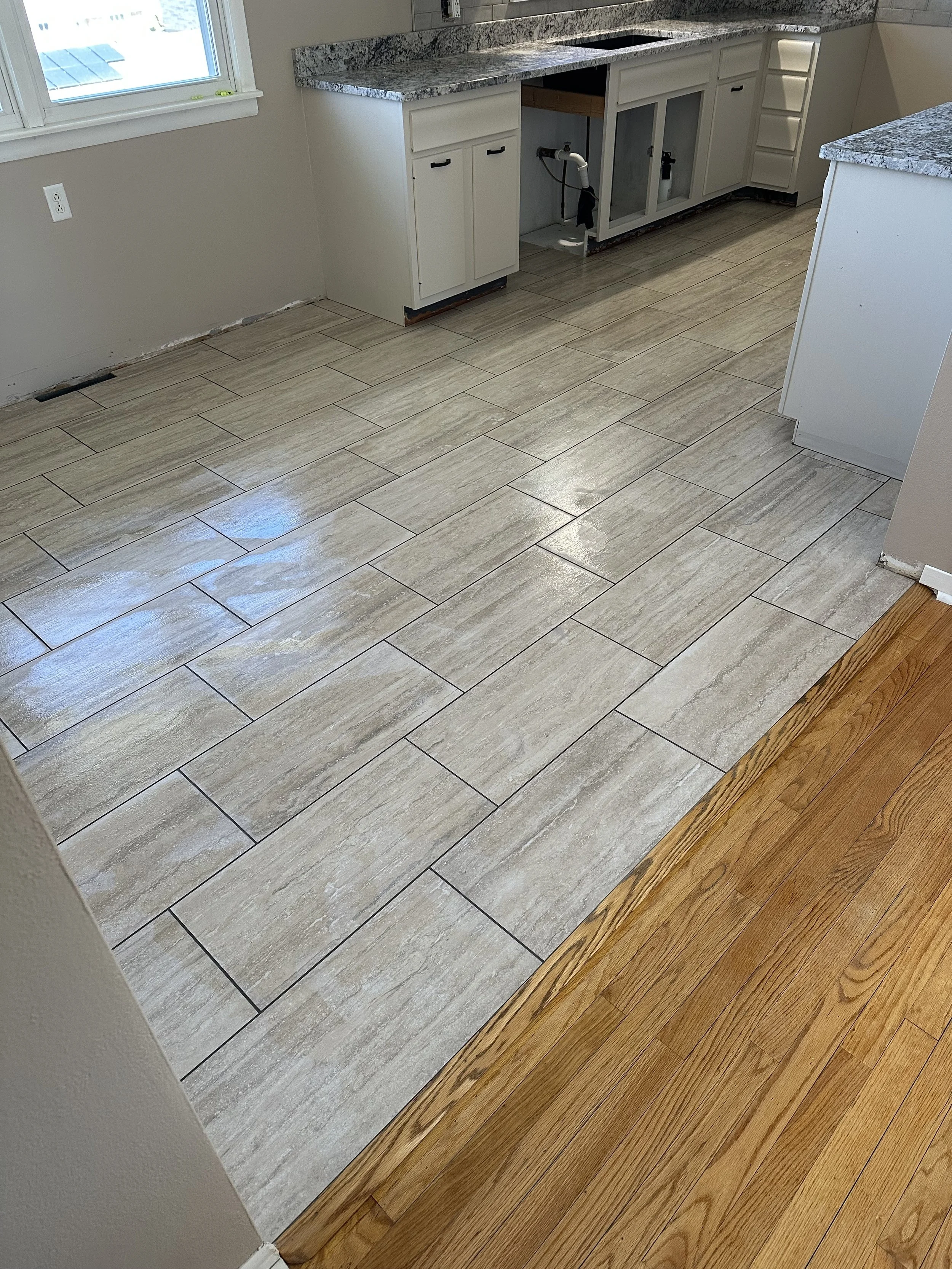 Image of a kitchen under renovation showing a contrast between tile flooring and hardwood flooring, with cabinets and a granite countertop, and an open space under the counter where a dishwasher or appliance was removed.