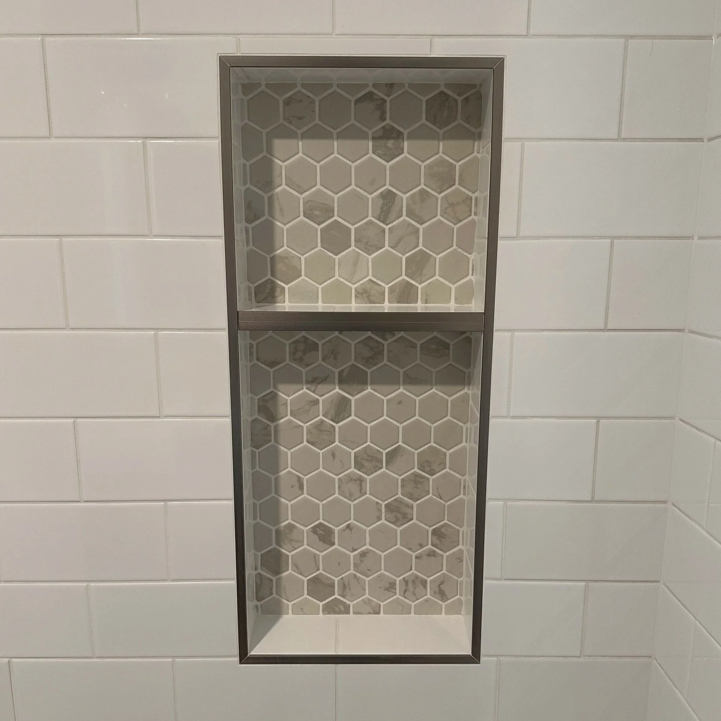 A recessed shower niche with a hexagon tile pattern and a metal frame installed in a bathroom wall with white subway tiles.