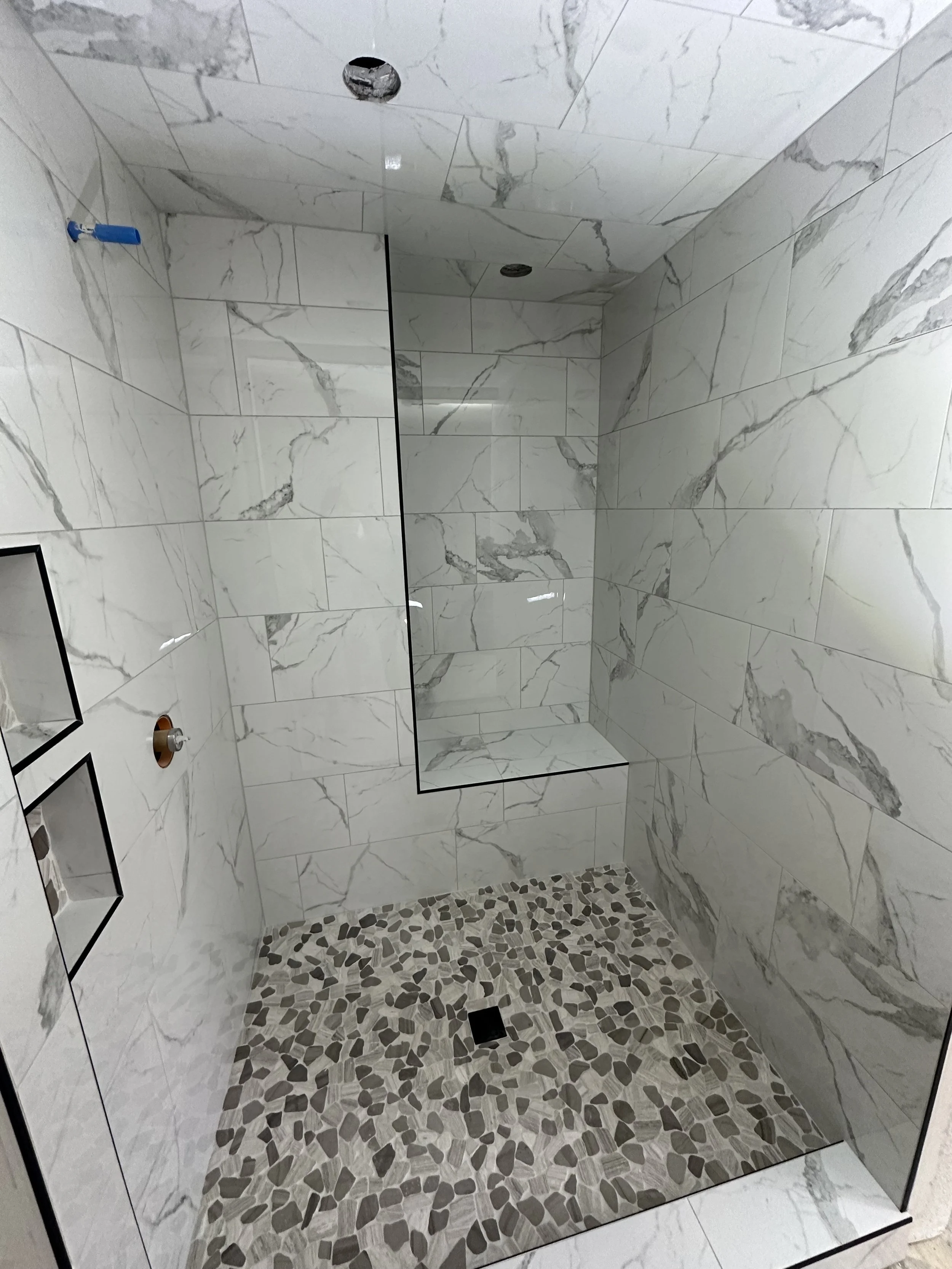 A modern walk-in shower with marble tiles on the walls and a pebble stone floor. There are two ceiling-mounted shower heads and built-in shelving.