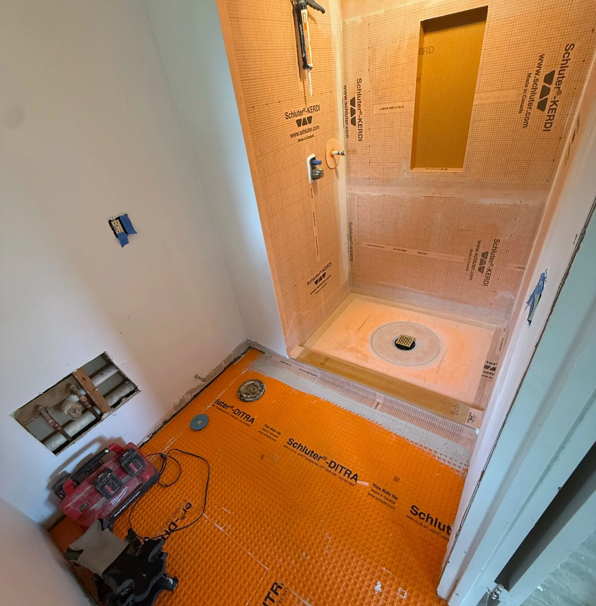 Bathroom under construction with a bathtub and plumbing exposed. Orange insulation and protective sheeting on the walls, tools on the floor, and an open box in the wall cavity.