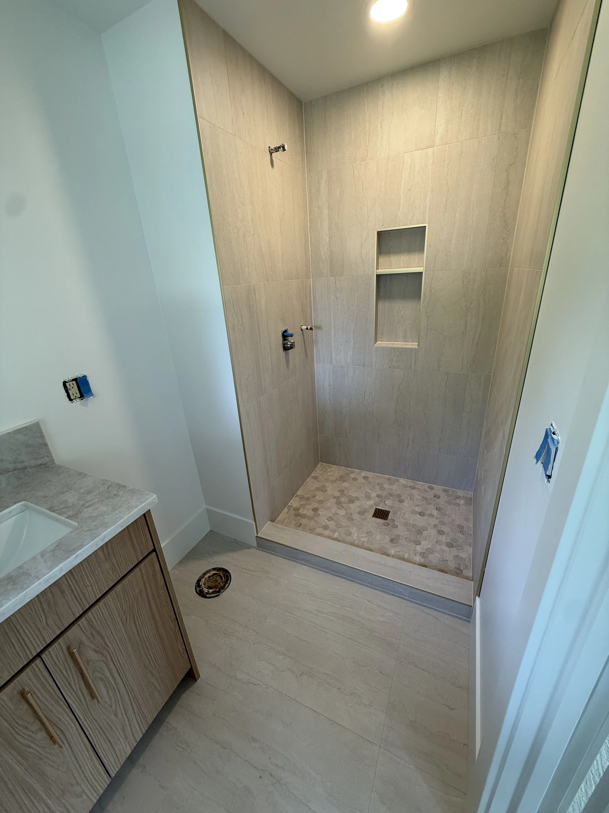 A bathroom shower area under construction with beige tiles, a recessed shelf, and an unfinished plumbing fixture. The image shows the floor drain and a nearby wooden cabinet with a marble countertop.