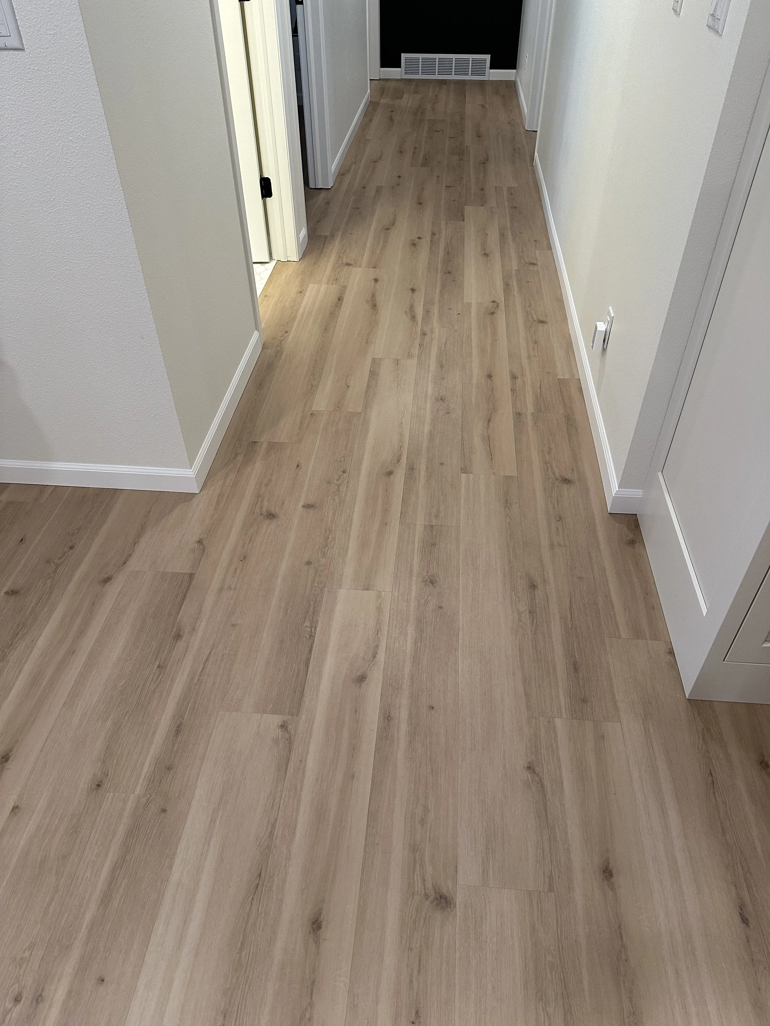 A hallway with light-colored wood flooring, white walls, and white door trim. There is a vent at the end of the hallway and two closed doors on either side.