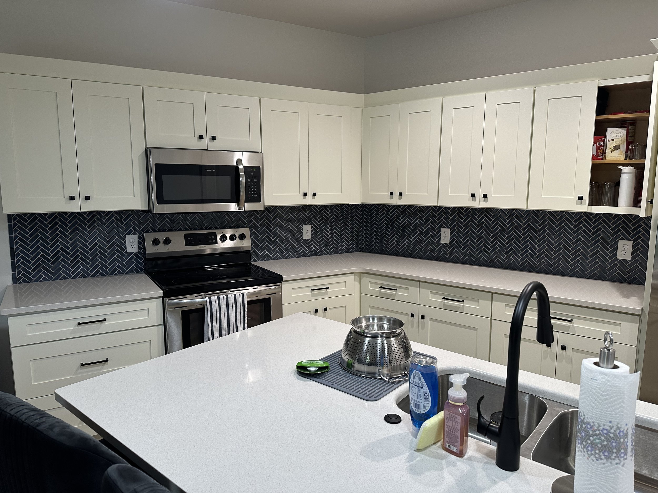 Modern kitchen with white cabinets, black hardware, stainless steel microwave, black stove, black and white backsplash, white countertops, a sink with a black faucet, items on the counter including hand soap, dish soap, a yellow sponge, a stainless steel strainer, and paper towels.