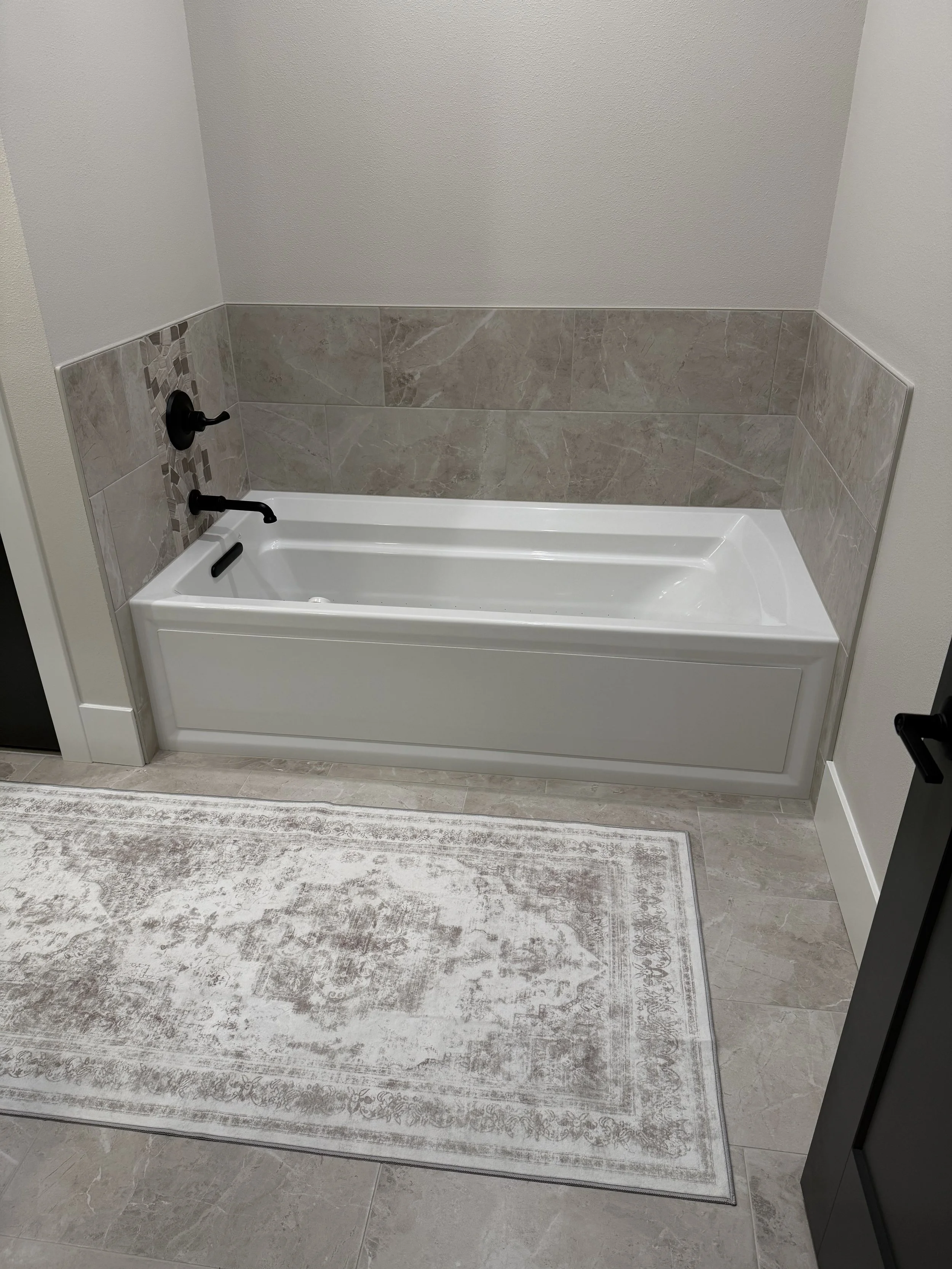 Bathroom with gray tiled walls, a white bathtub, black fixtures, a beige and cream rug on gray tile floor.