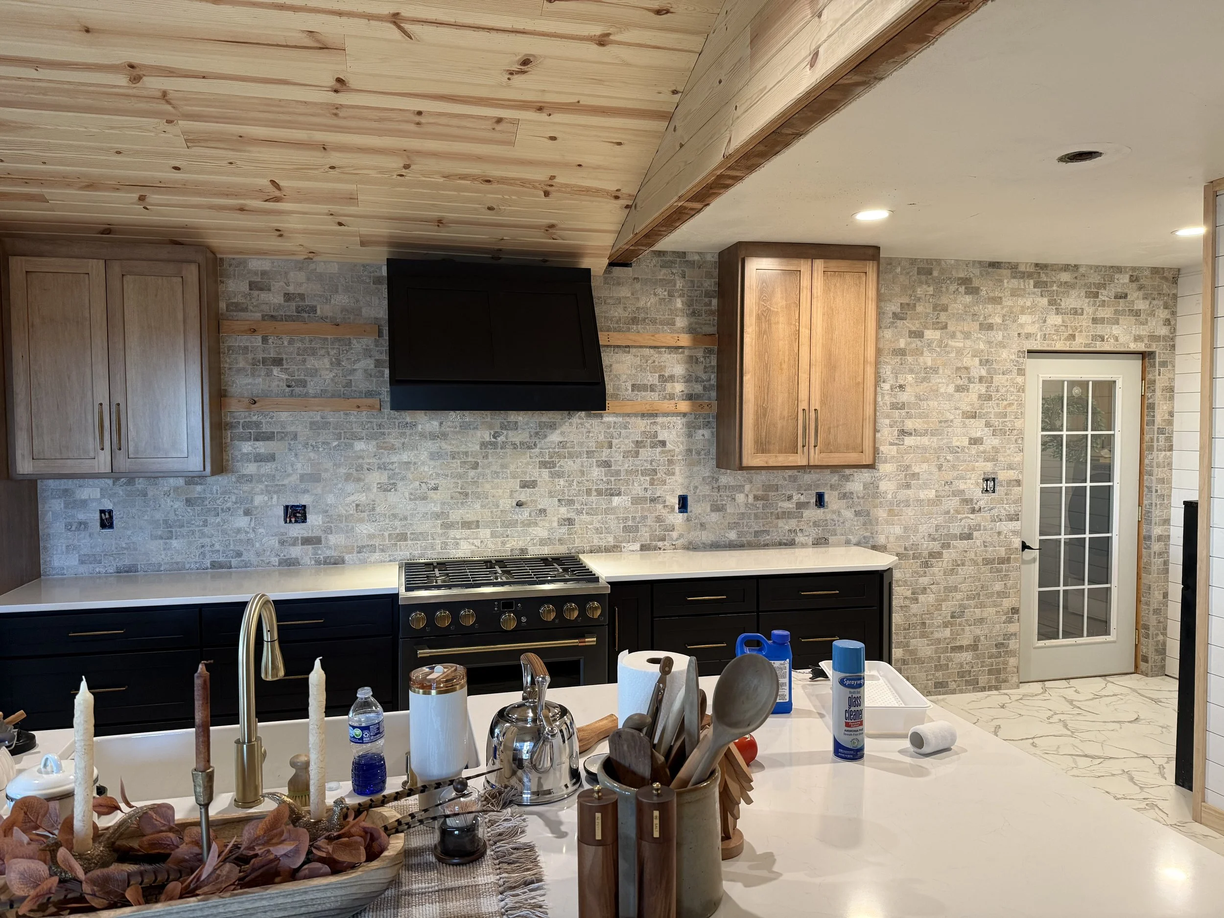 A kitchen with a wooden ceiling, brick walls, and cabinetry. Various items are on the white countertop, including soap, cleaning spray, and utensils. A stove and a window are visible.