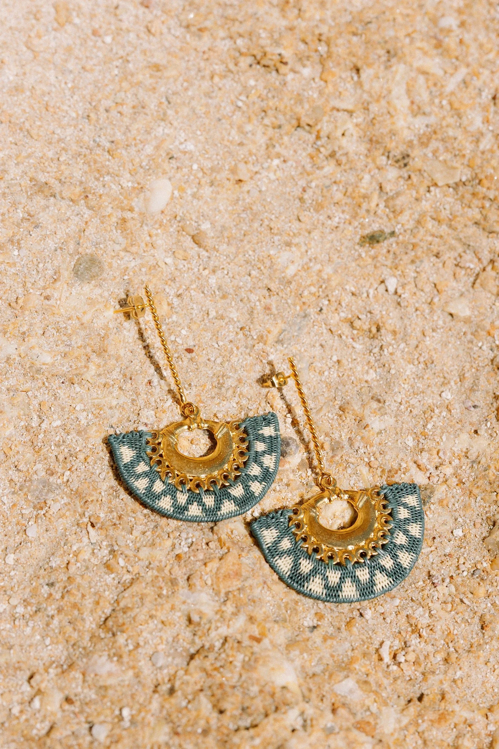 Pendientes de aro dorados con tela en color azul y blanco, colocados sobre arena.