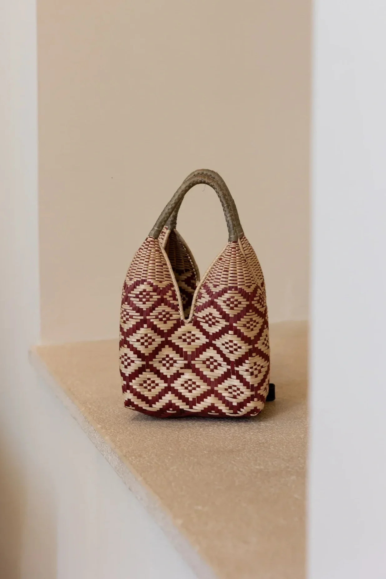 Bolso tejido con patrón de rombos en tonos marrón, beige y rojo, con asas cortas de cuerda de yute, colocado sobre una repisa beige.