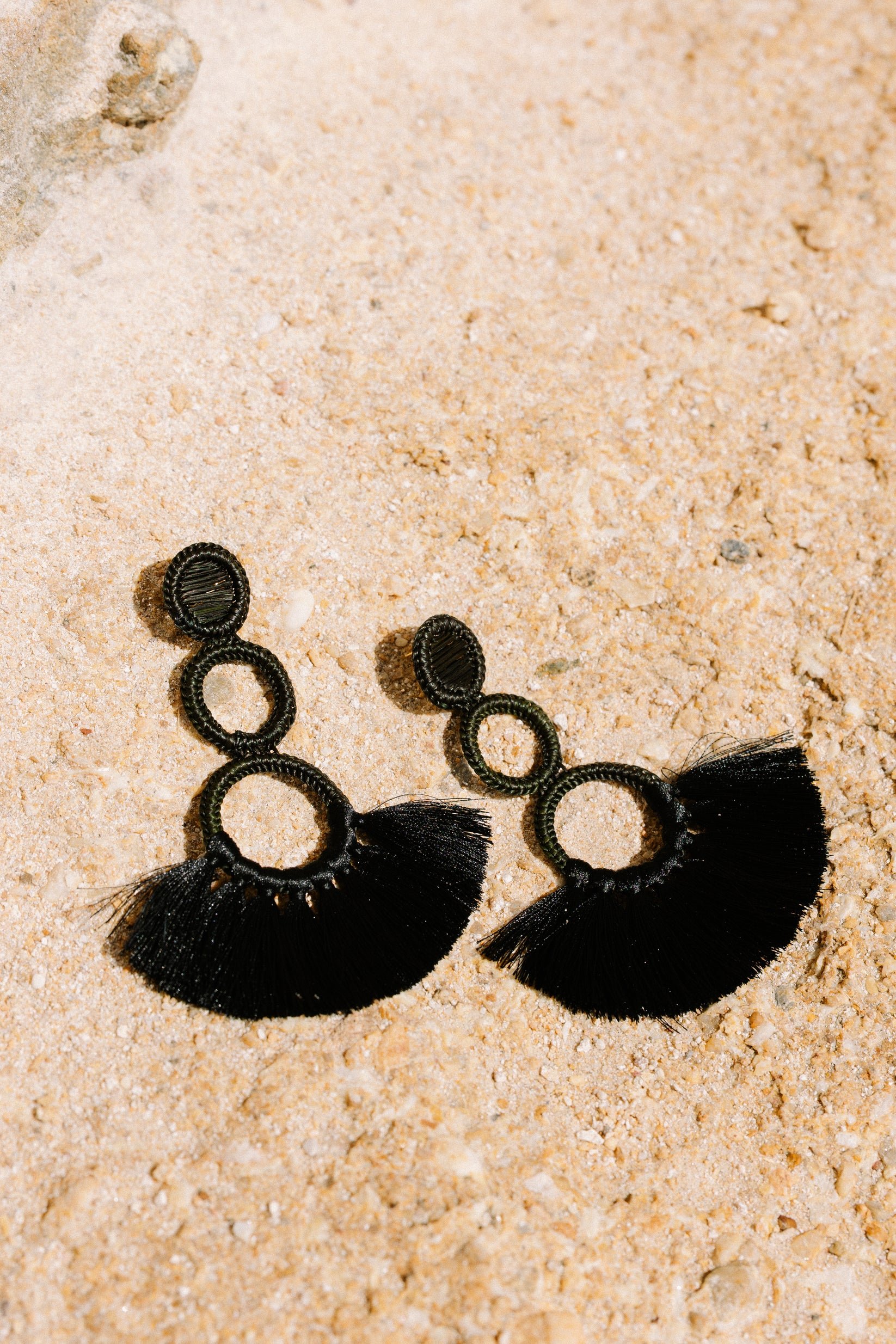 Pendientes negros con flecos, colocados en arena clara.