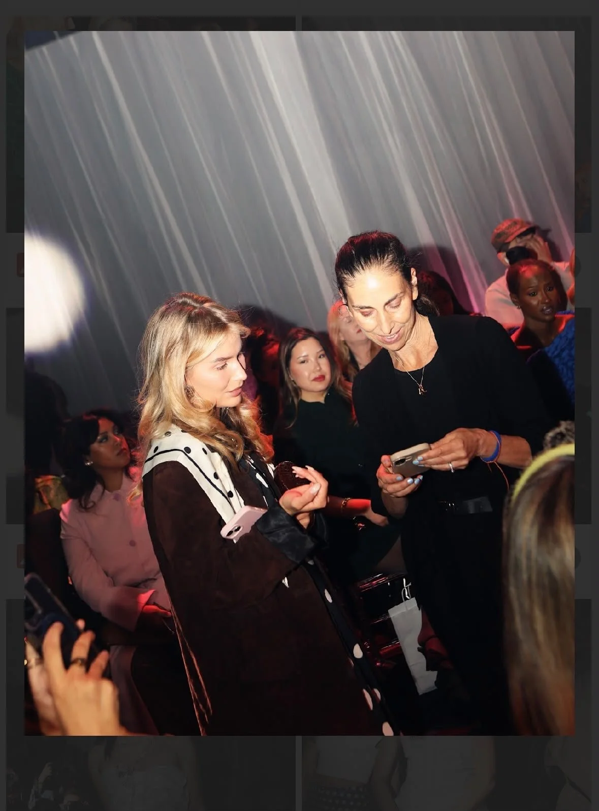 Mujeres en un evento social, mirando teléfonos móviles, en un ambiente con iluminación suave y cortinas blancas en el fondo.