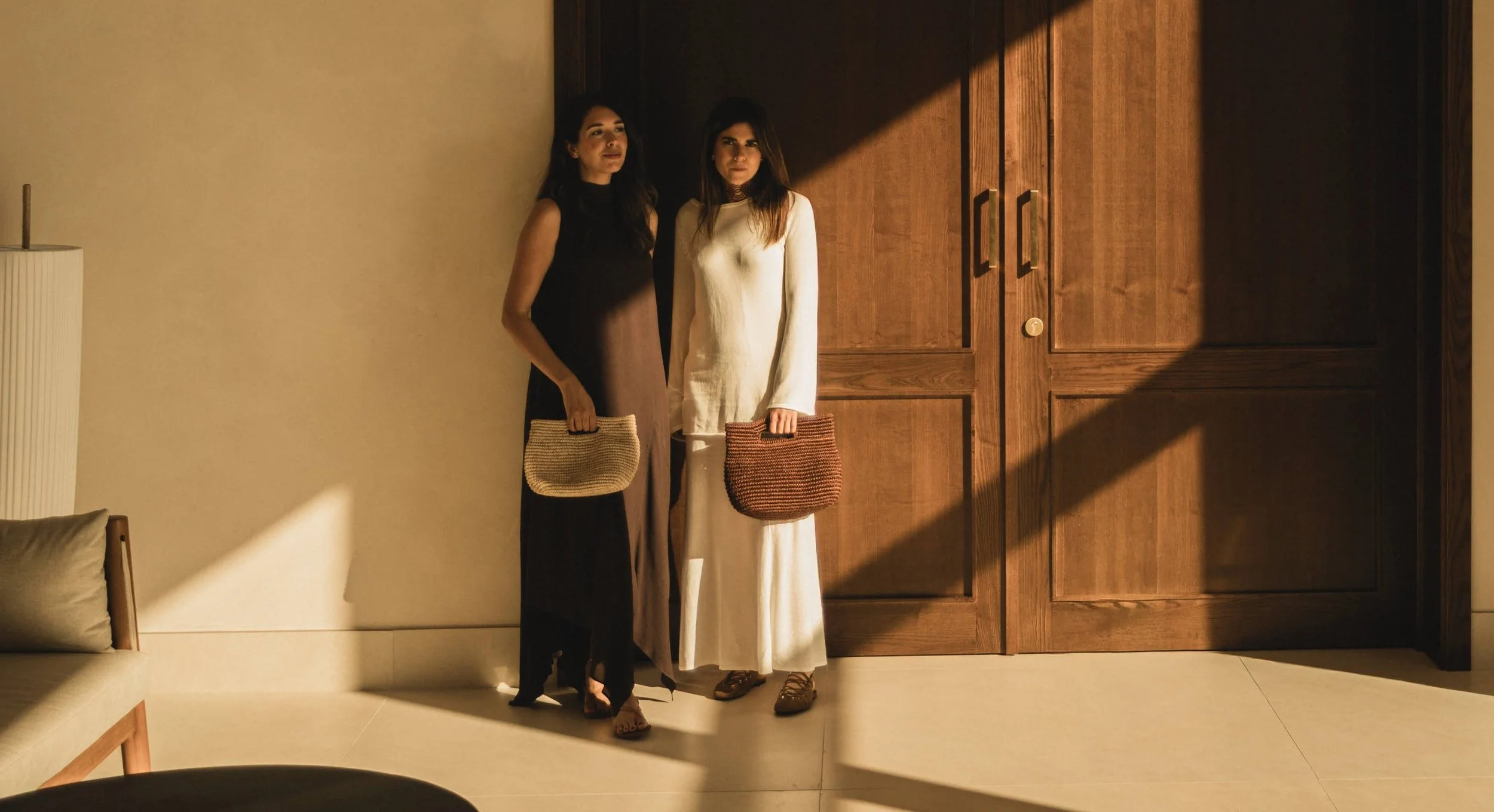 Dos mujeres de pie sosteniendo bolsas de mano en una sala con puertas de madera y luz que entra por una ventana, creando sombras en la pared.
